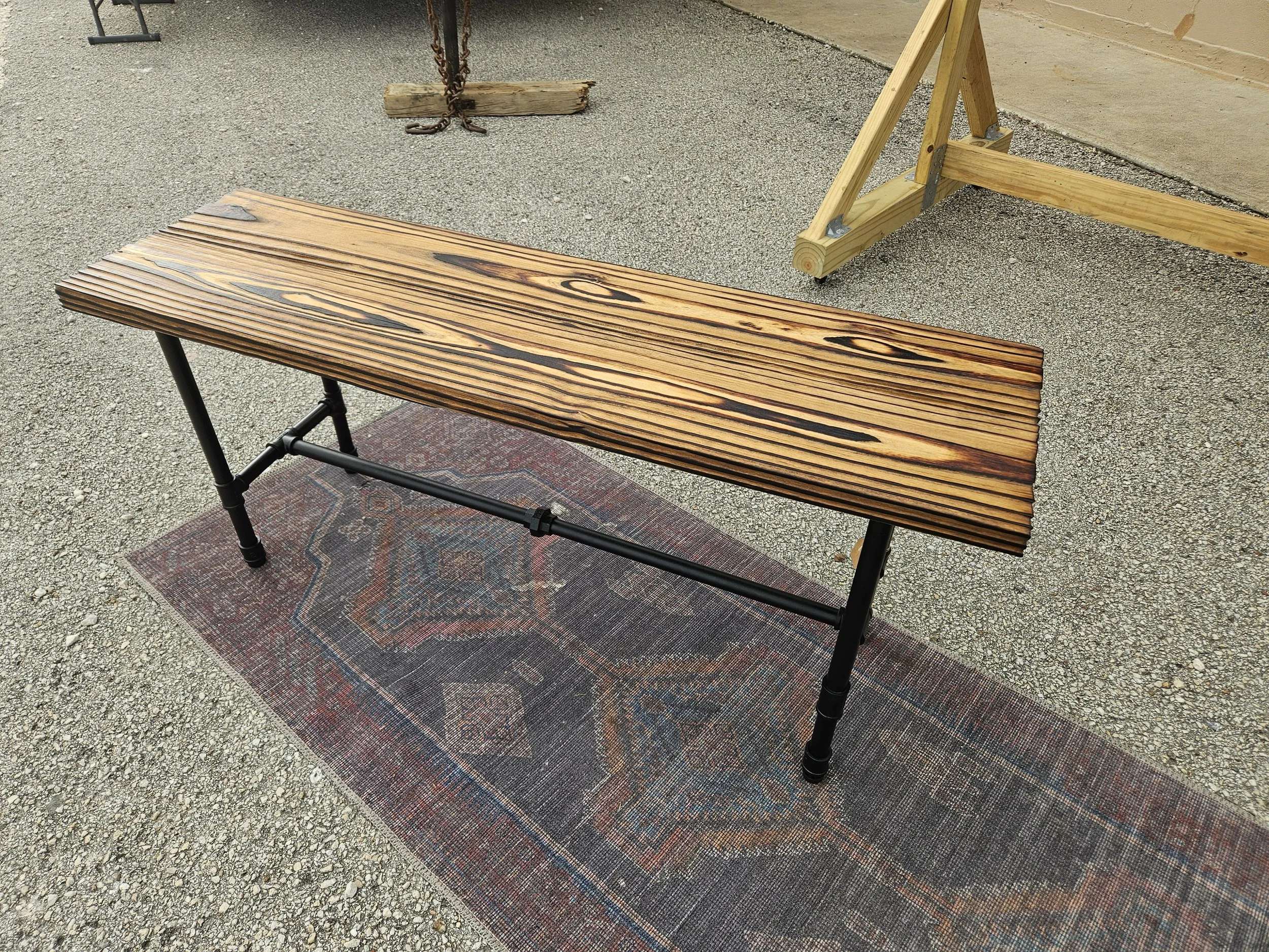 A wooden bench with a dark finish is placed on a patterned rug on top of a gravel surface. Next to the bench, there is a wooden sawhorse with metal brackets. In the background, there is a wooden beam supported by chains and a wooden block, as well as