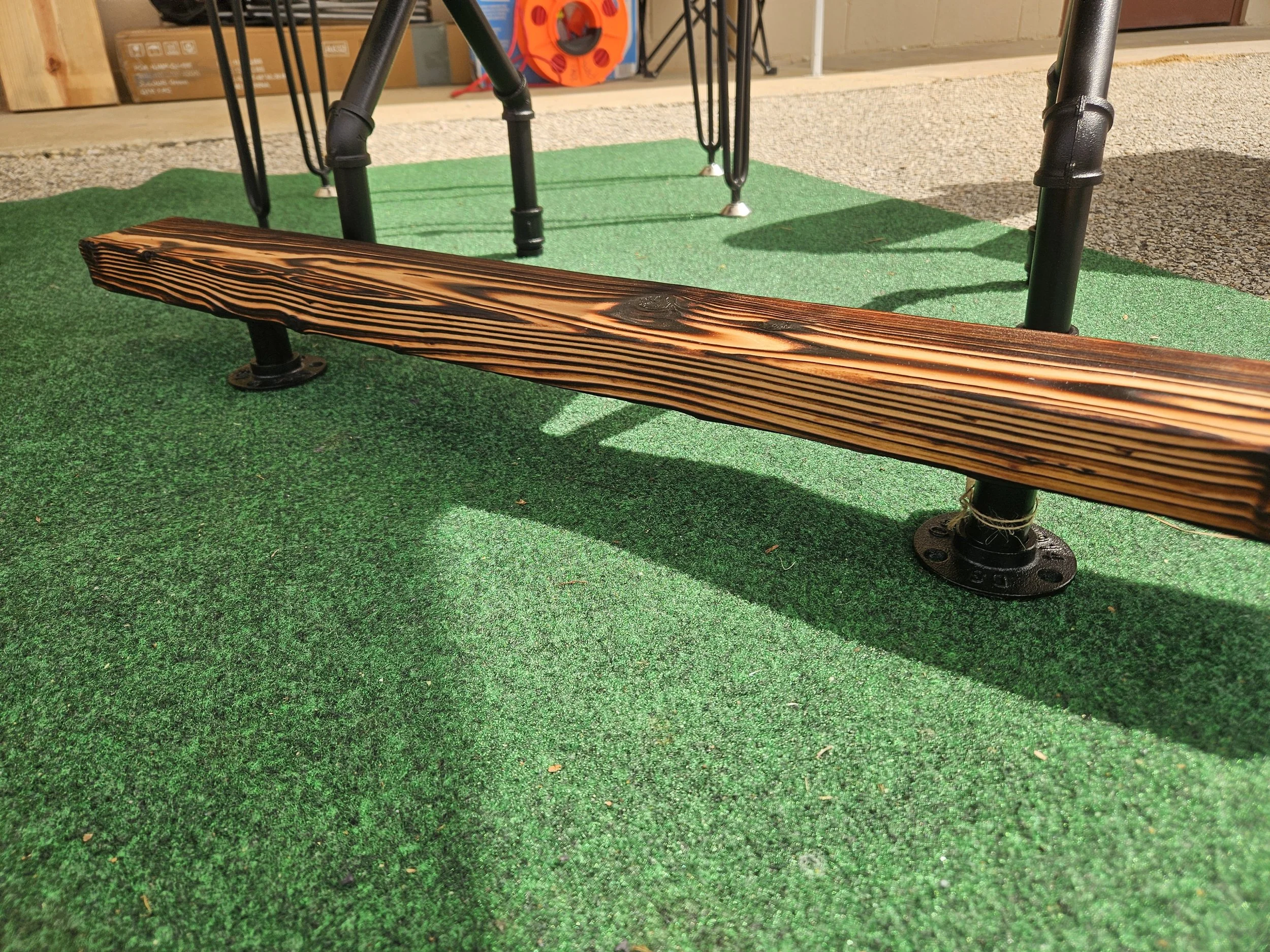 Close-up view of a wooden bench with black metal legs on an outdoor green artificial turf surface, in front of a paved area and building with cardboard boxes and storage items in the background.