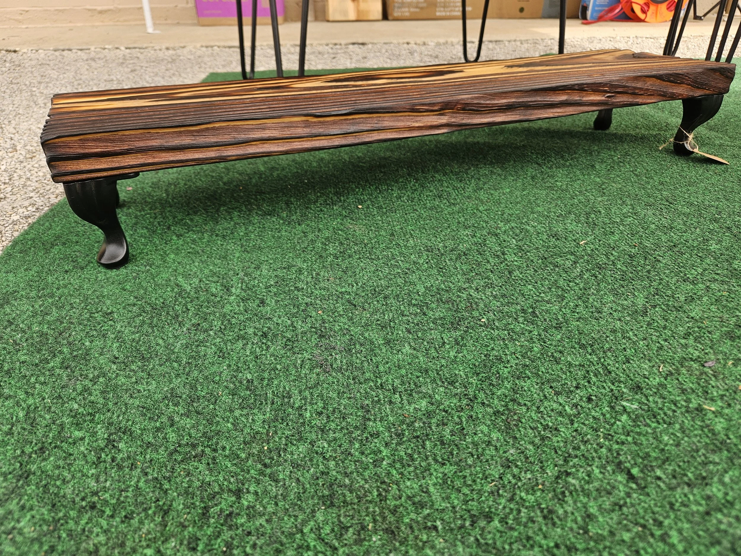 Long wooden bench with curved black metal legs on green artificial grass, with a gravel pathway and store background.