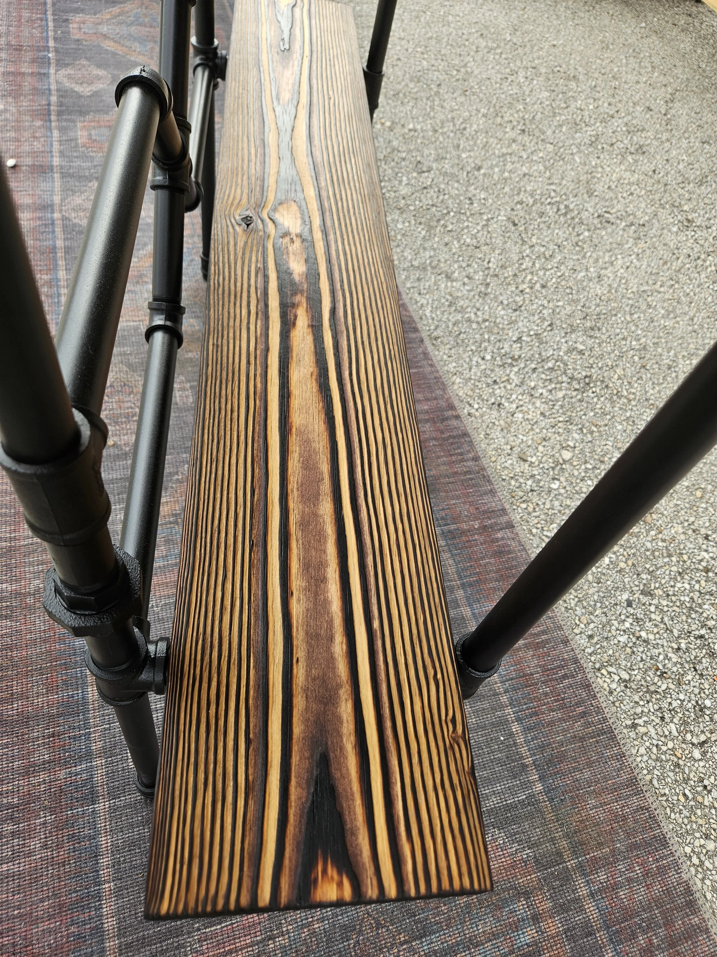 A wooden bench with a dark natural finish, supported by black metal legs, placed on a decorative outdoor rug with geometric patterns, next to a concrete sidewalk.