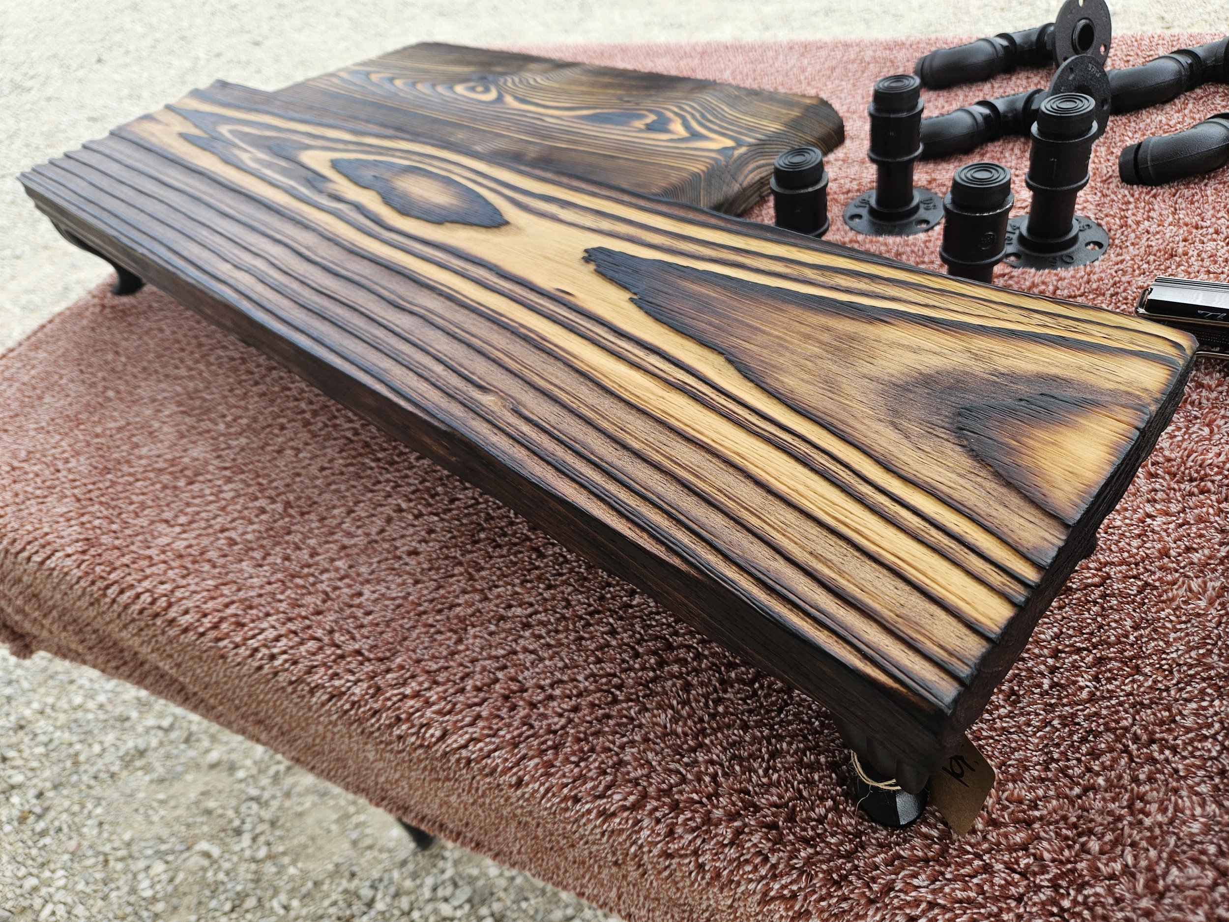 Close-up view of a wooden table with a burnt or charred wood finish and black pipe fittings on a pink textured cloth, with a gravel surface in the background.