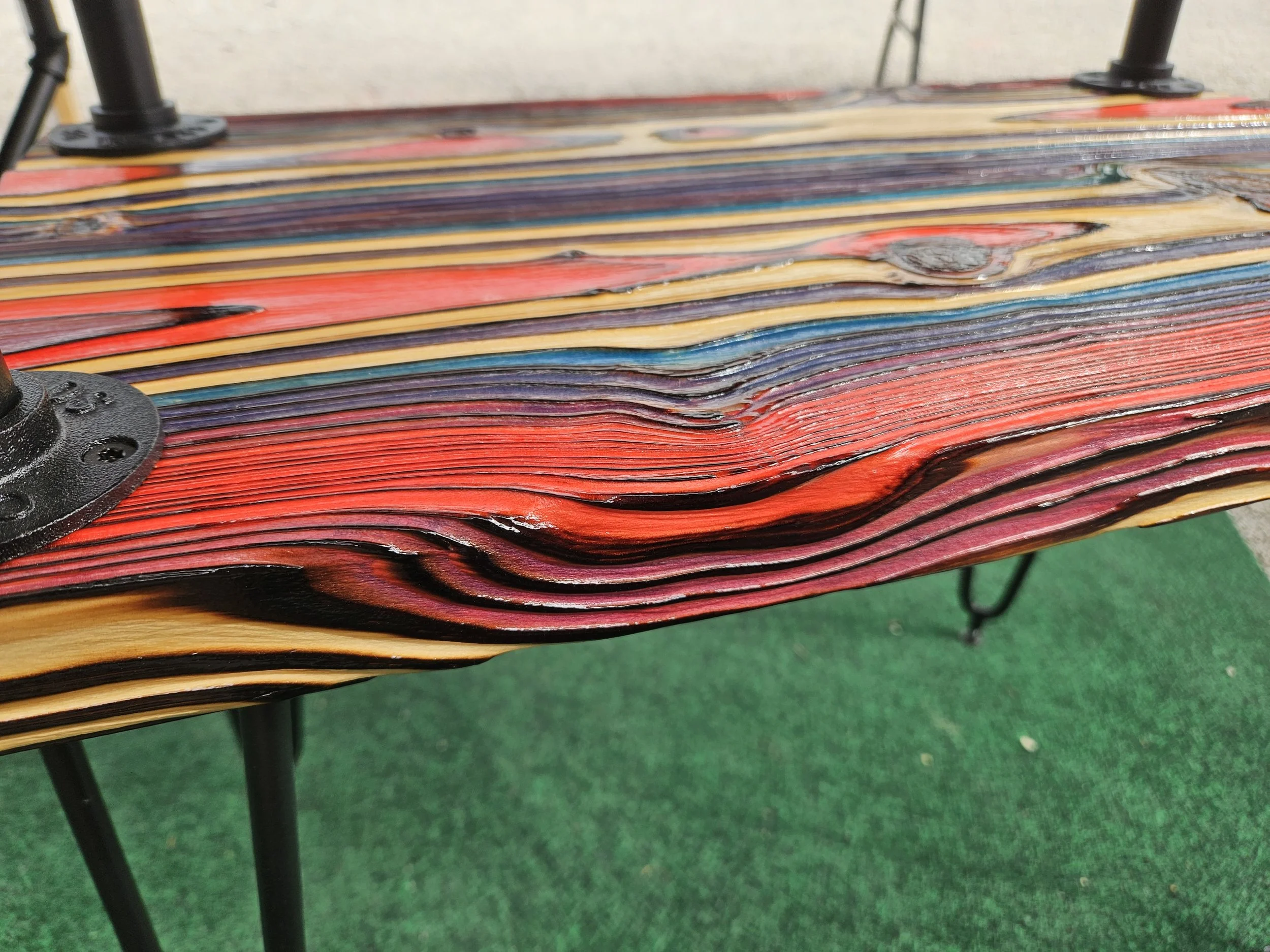 Close-up of a multicolored wooden bench with black metal supports in an outdoor setting with green ground surface.