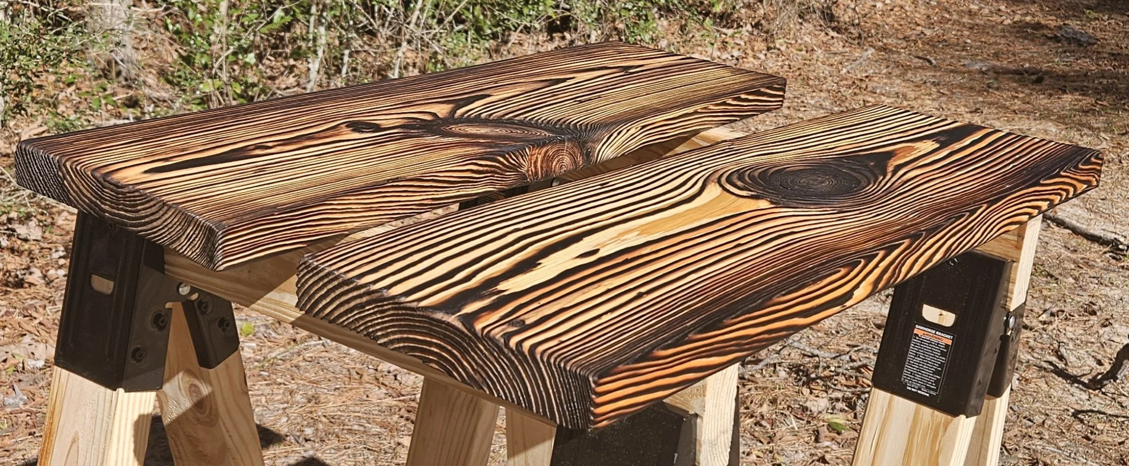 A handmade wooden picnic table outdoors, with burnt wood finish on the surface, set on a dirt ground with leaves and greenery in the background.