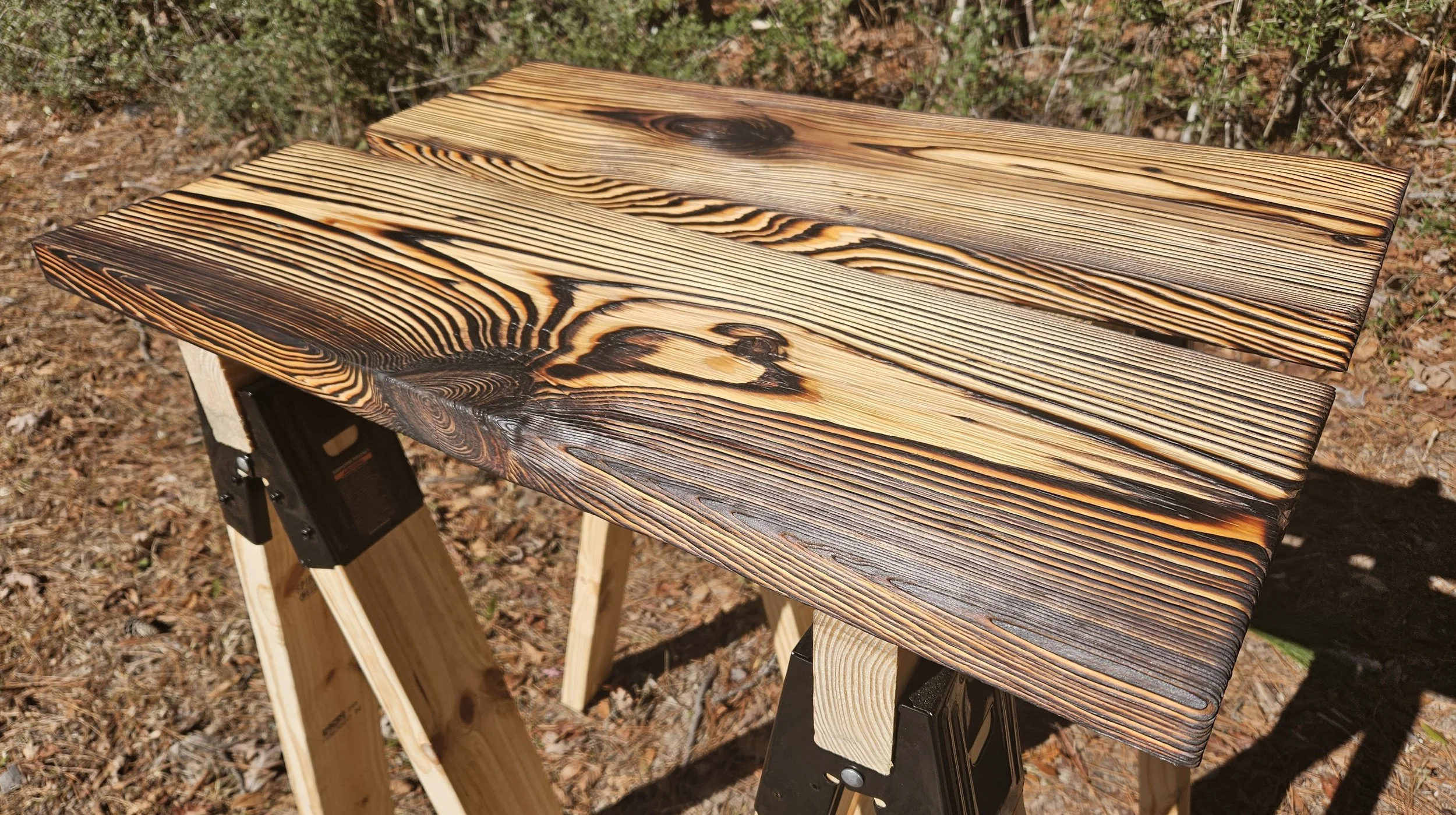 Two pieces of wood with visible grain patterns and some burn marks resting on sawhorses outdoors.