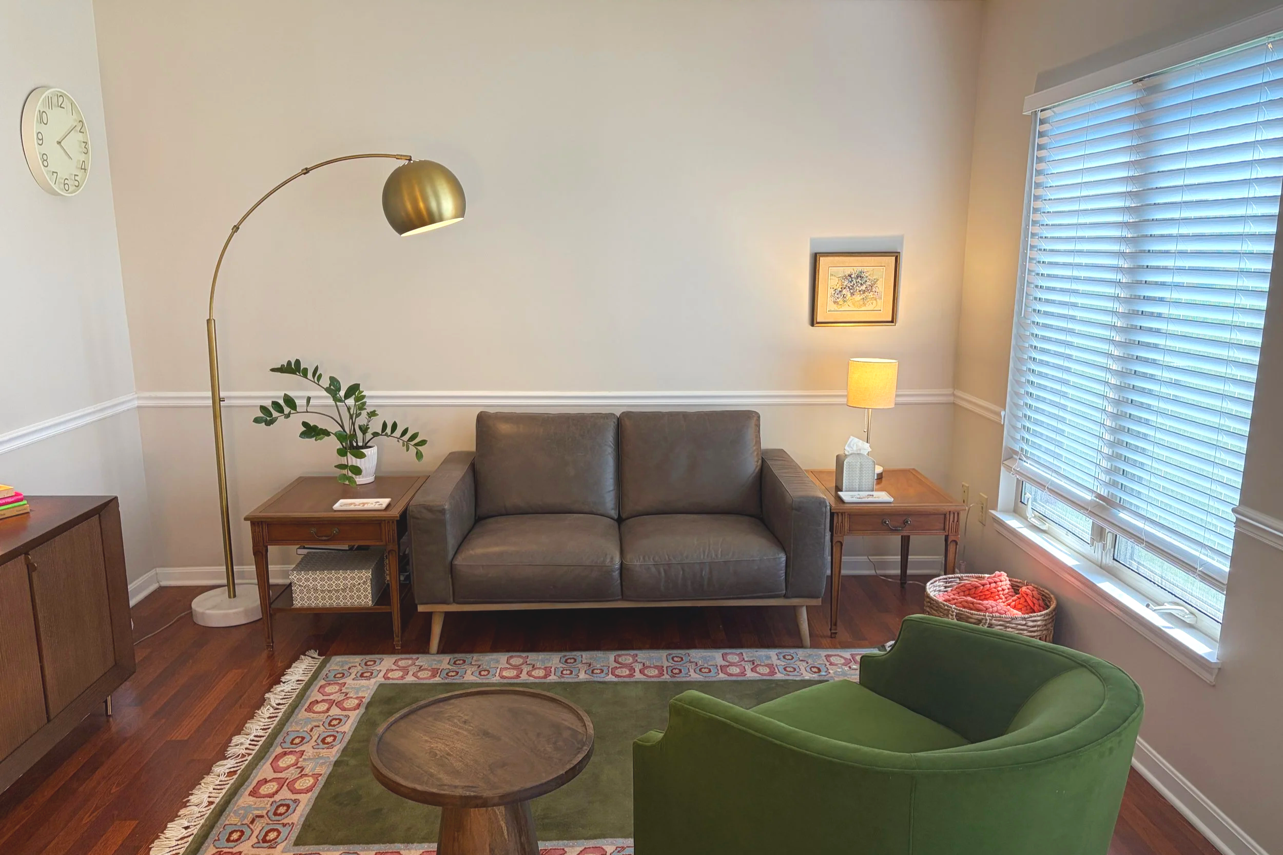 Living room with a dark gray couch, a green armchair, a wooden coffee table, a patterned rug, a tall floor lamp, a side table with a plant, a framed picture, and a large window with blinds.