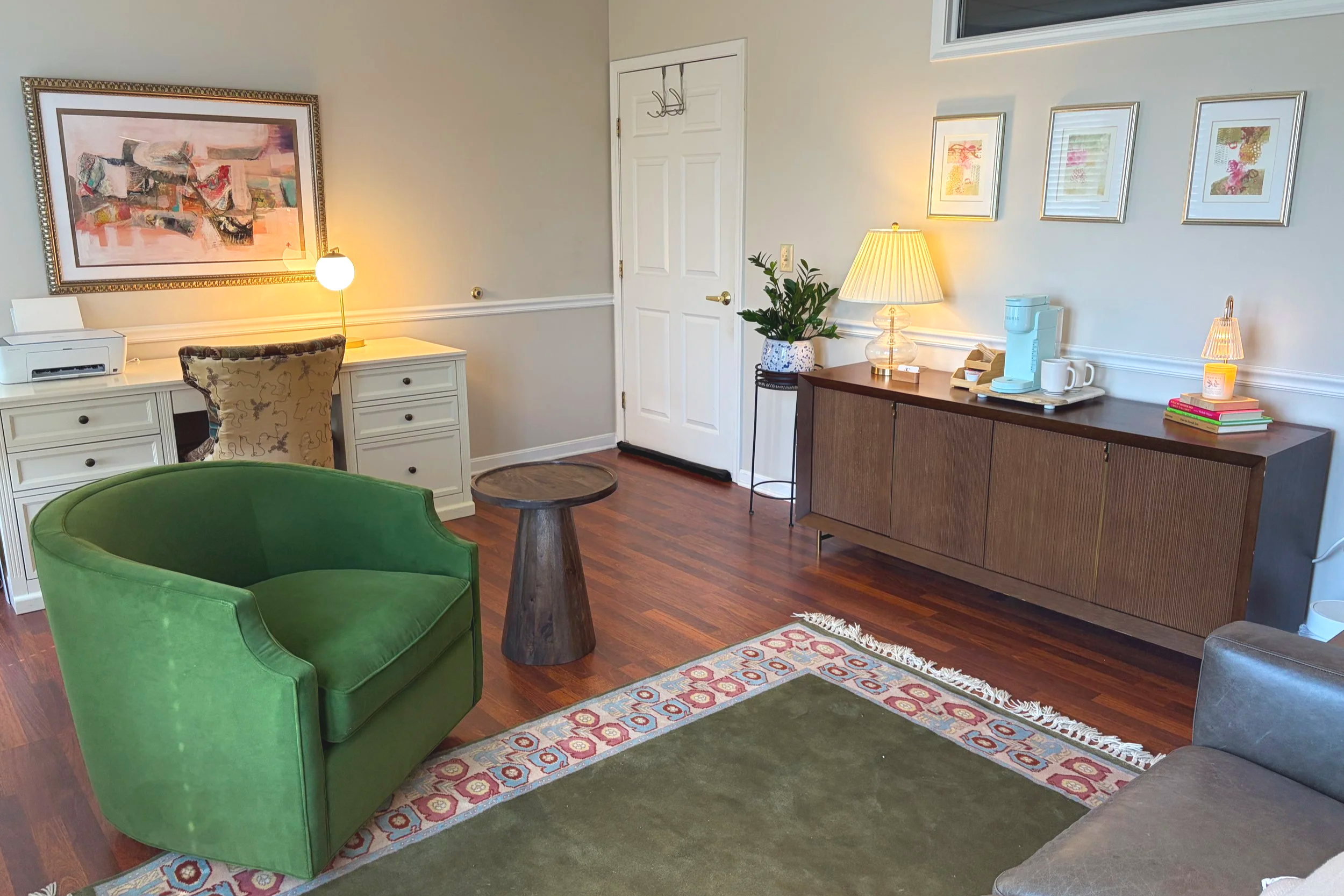 Interior of a cozy living room with a green armchair, a wooden side table, a white desk with a printer, a potted plant on a black stand, a buffet with a lamp, a coffee maker, cups, books, and framed artwork on the wall.