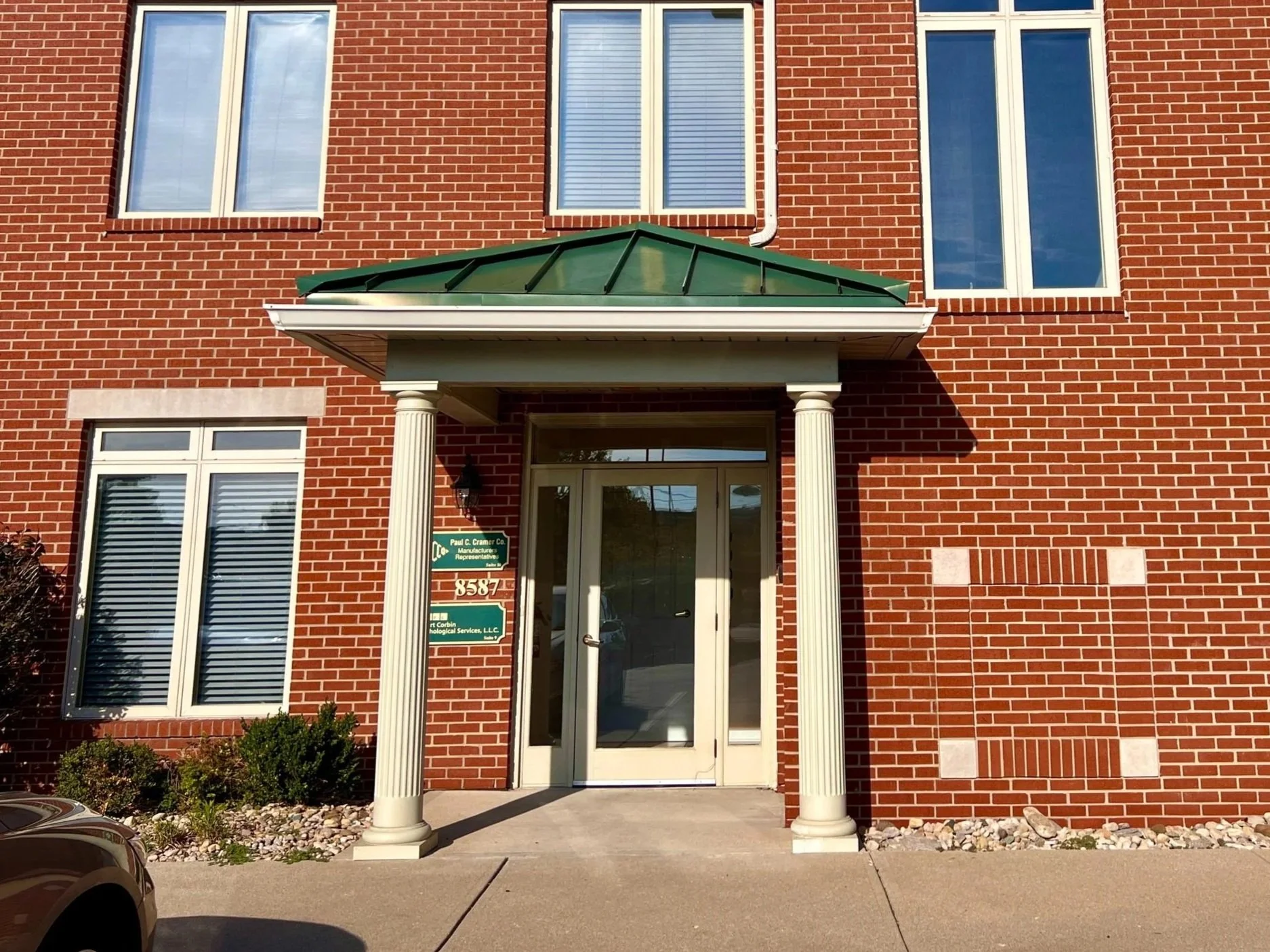 Front view of a red brick commercial building with two stories, featuring three tall windows with white trim on the upper level, a glass door entrance with a white frame, and decorative white columns supporting a green metal awning over the entrance.