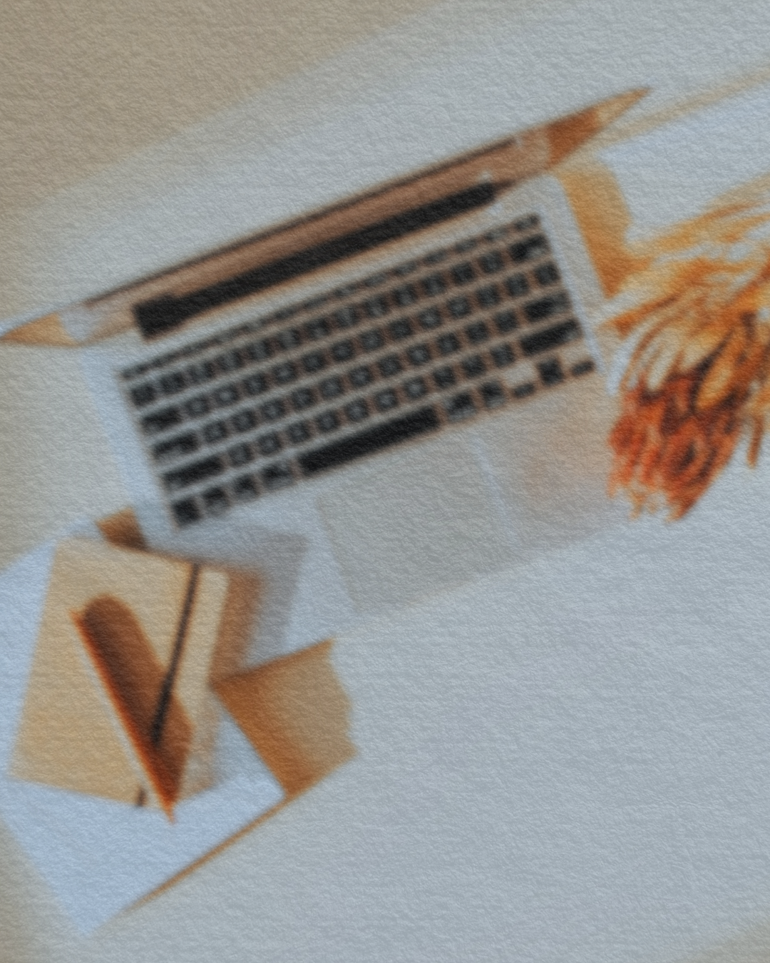 Blurry image of a laptop, a small notebook, and a bunch of orange foliage on a white surface.