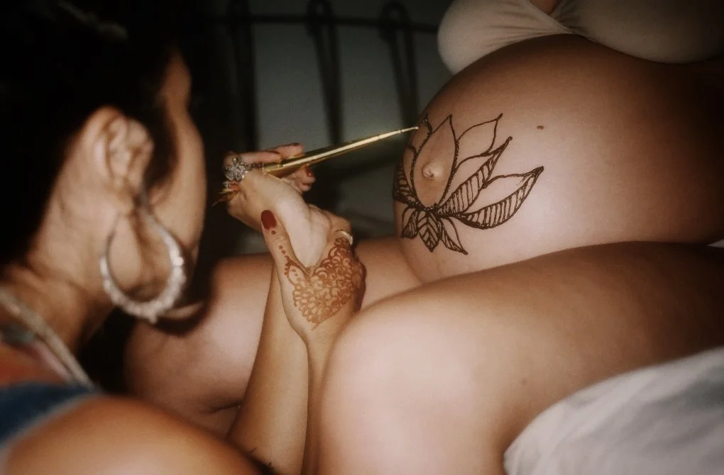 A woman with henna on her hand paints a lotus flower tattoo on another woman's pregnant belly using henna during a mother blessing also known as a blessing way.