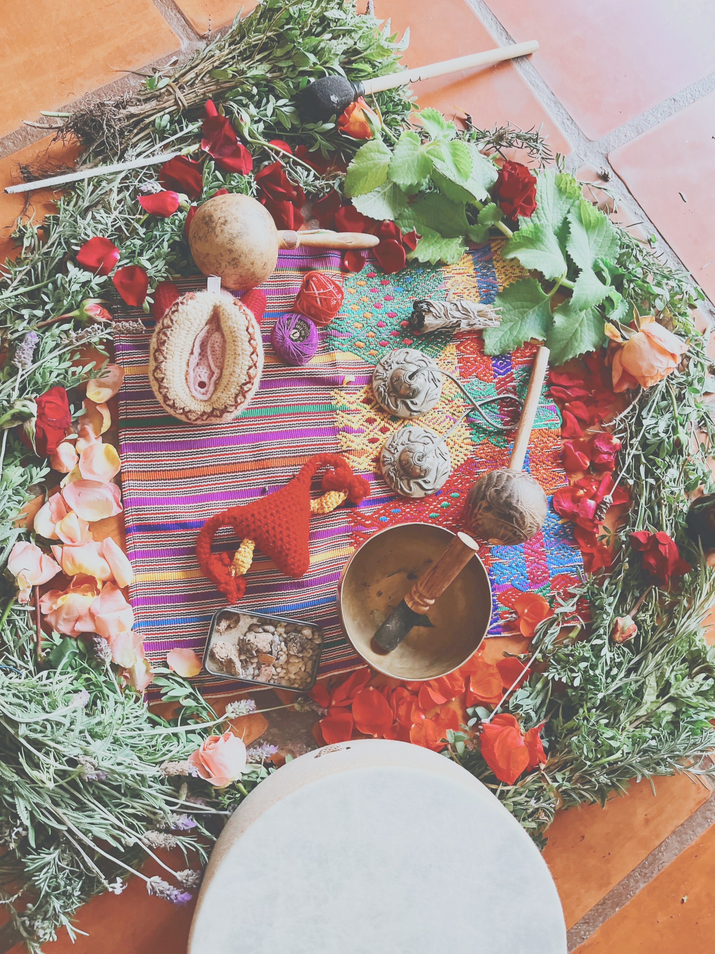 A colorful altar surrounded by flowers and greenery, with various objects including a bell, a crochet uterus, holy items, shells, and a sacred fabric.