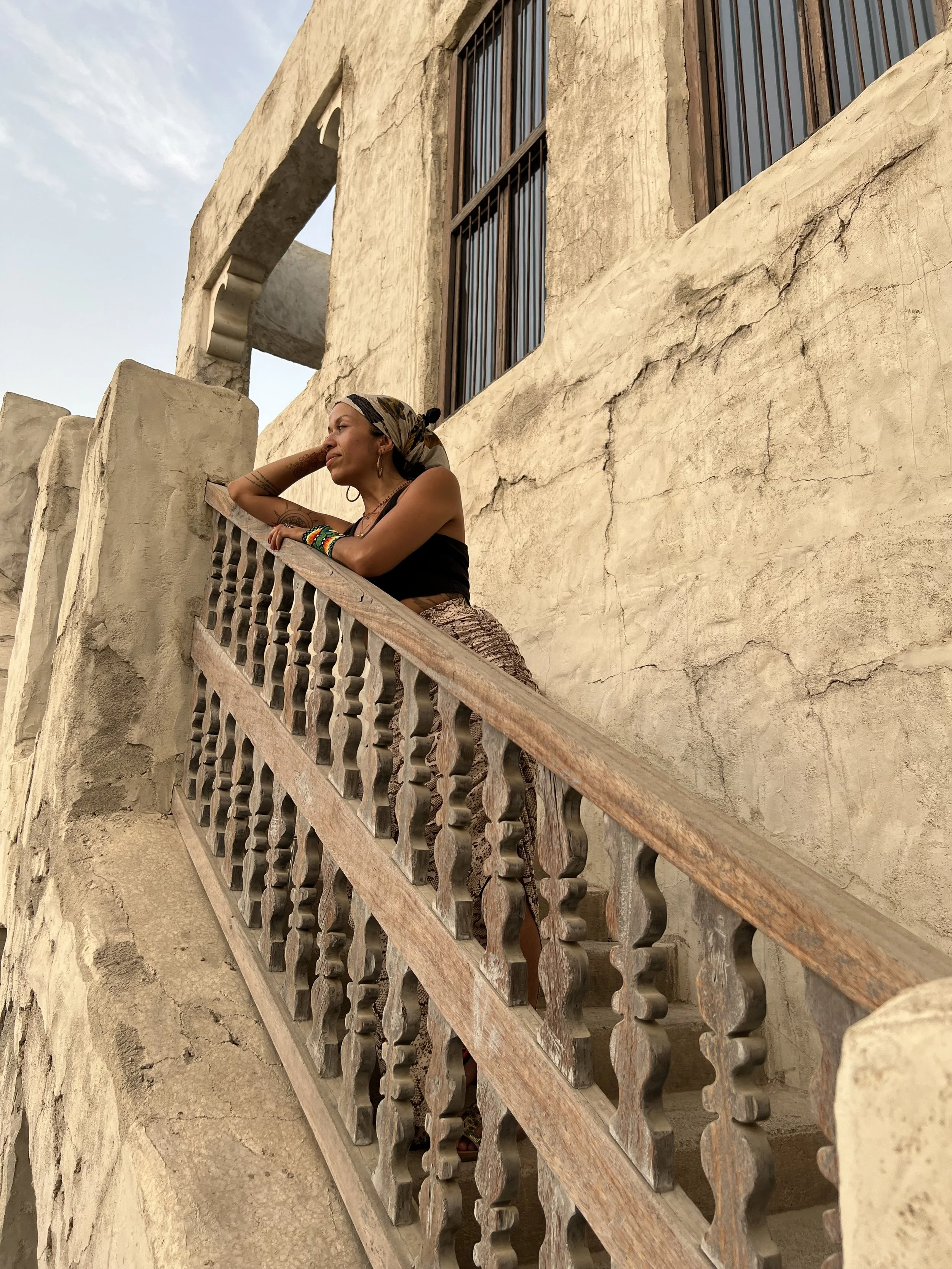 A shamanic woman, birthworker and doula leaning on a rustic wooden railing on a beige stucco building balcony, looking thoughtful as she gazes into the distance in Dubai