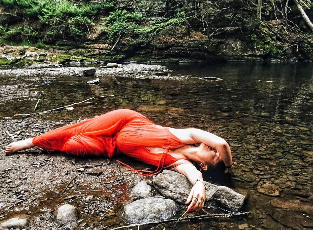 A pregnant woman in a red dress lying on her side on rocks by a calm, shallow river in a forested area during her maternity photo shoot by Vanessa Ixchel Fernandez