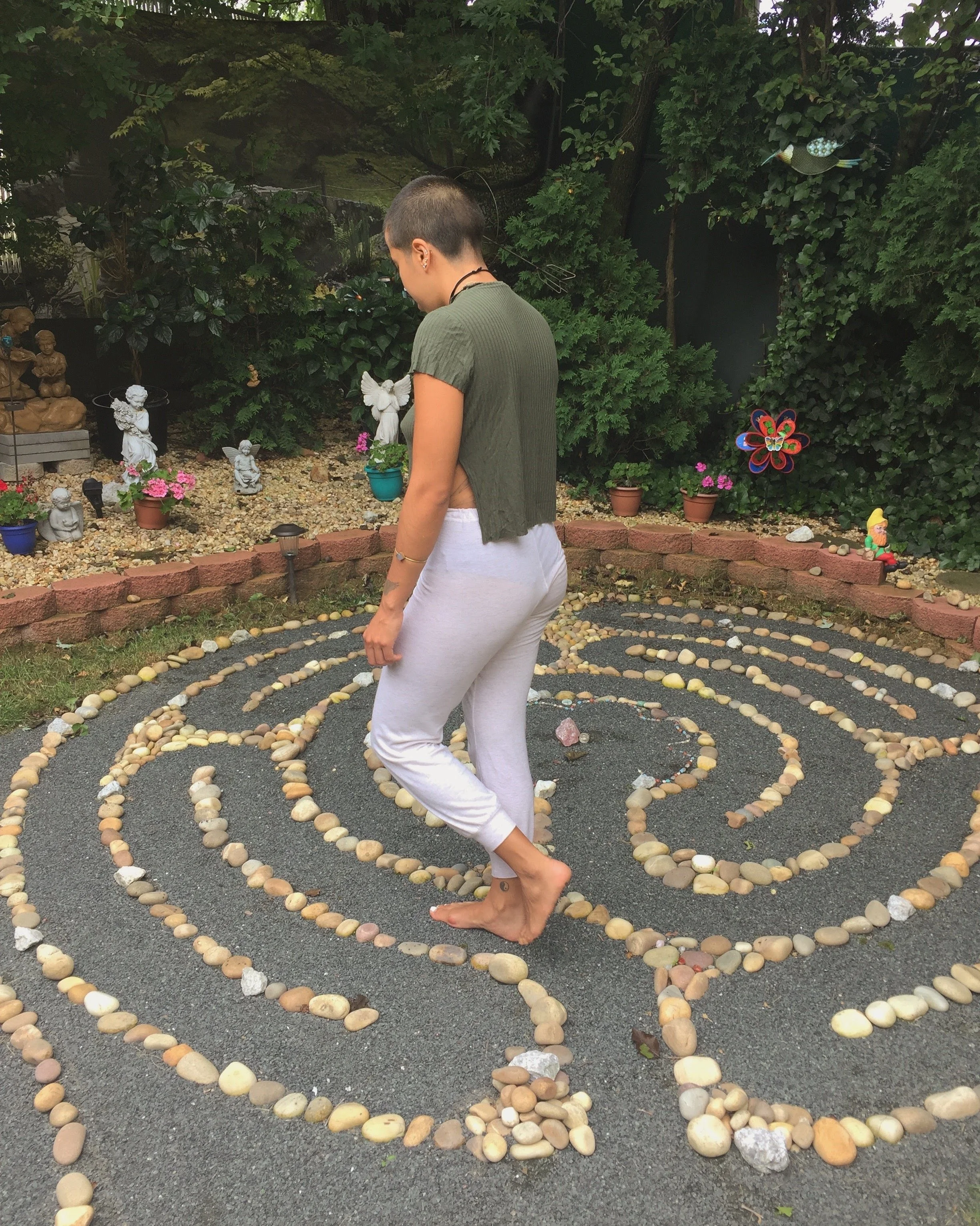 Person with short hair wearing a green top and white pants standing barefoot on a spiral-shaped pebble maze in a garden, surrounded by decorative plants and garden ornaments.