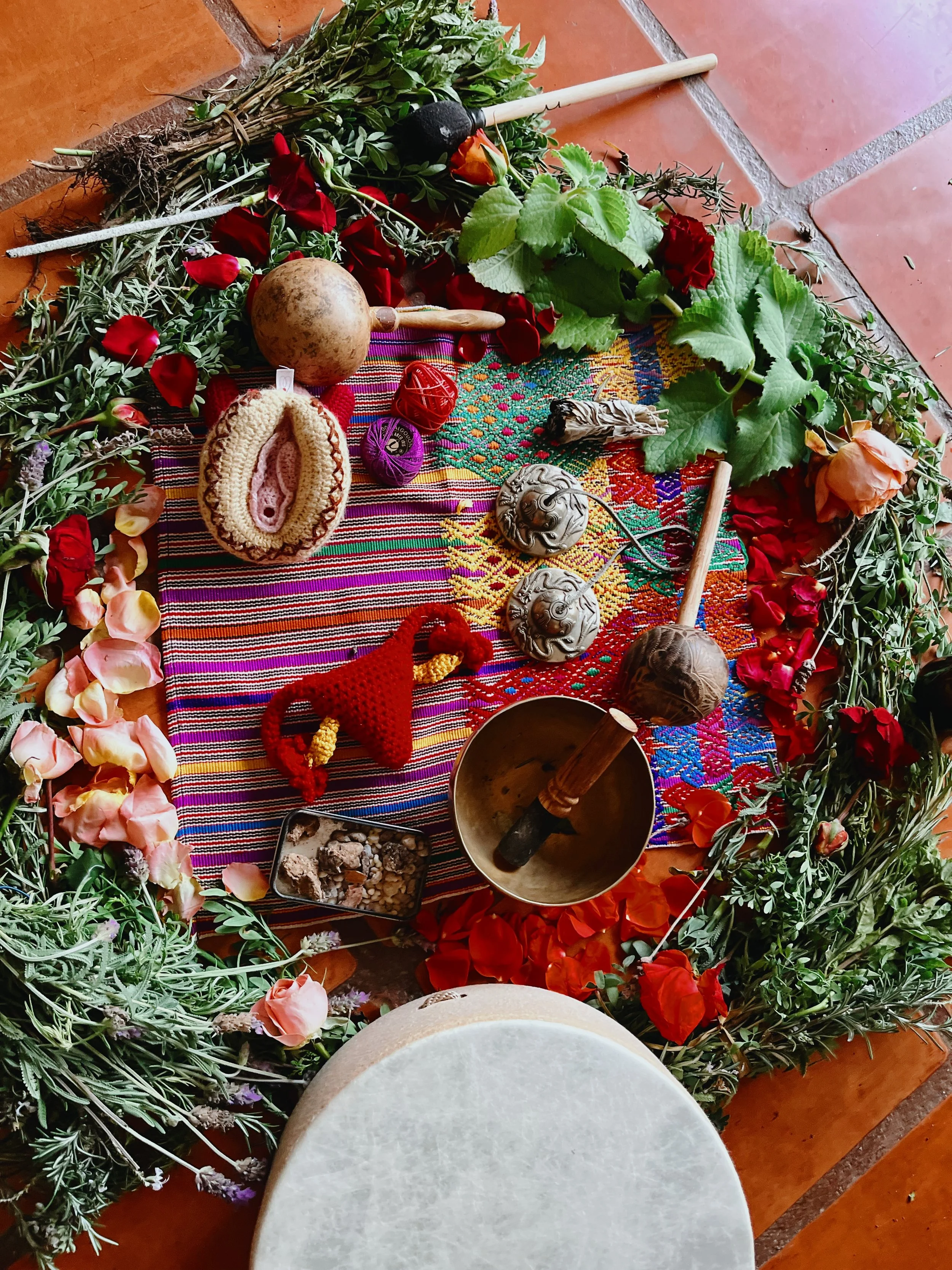 A sacred altar with a colorful woven cloth, surrounded by green foliage and flowers, containing various objects such as brass bells, a crocheted uterus, a small bowl with rocks, and a wooden shaker representing the ceremonial aspect of birth