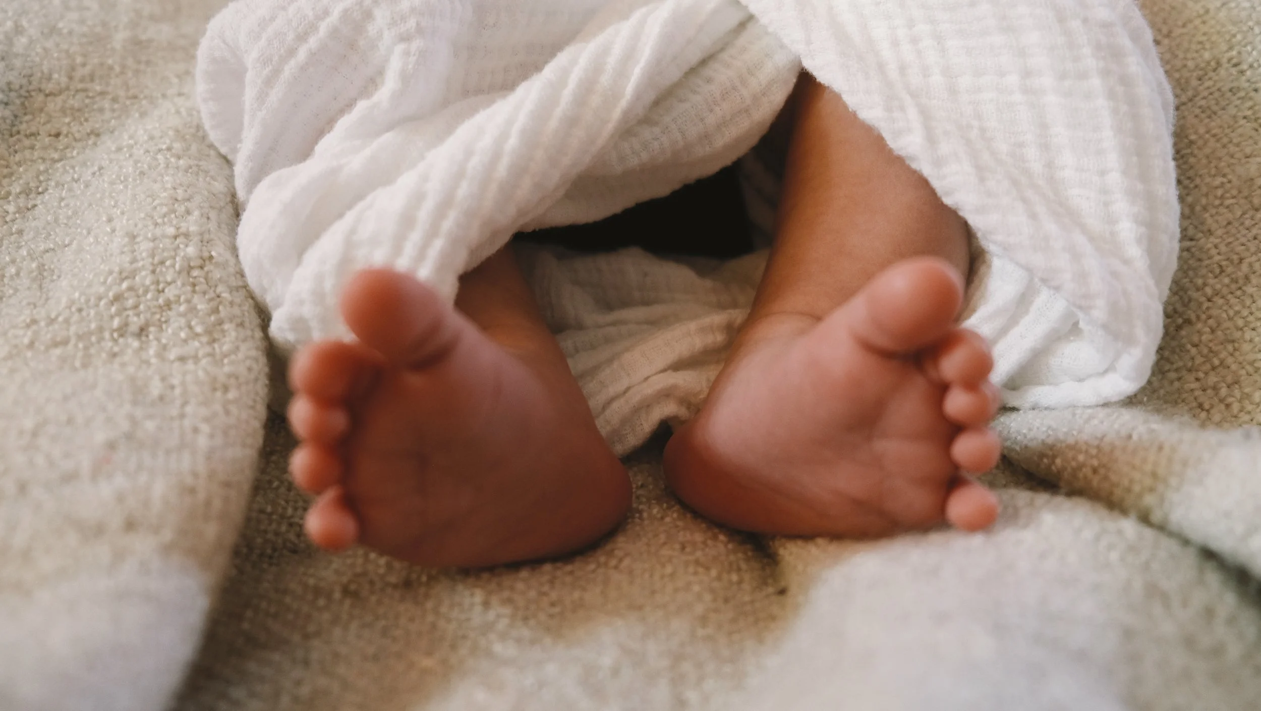 Close-up of a baby's feet with toes curled, partially wrapped in a white towel, resting on a soft beige fabric surface. Postpartum photoshoot by Vanessa Ixchel Fernandez