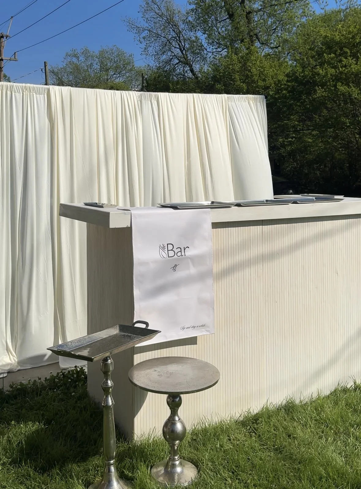 A white wooden bar in front of a white linen back-drop. There are two small silver tables off to the side.