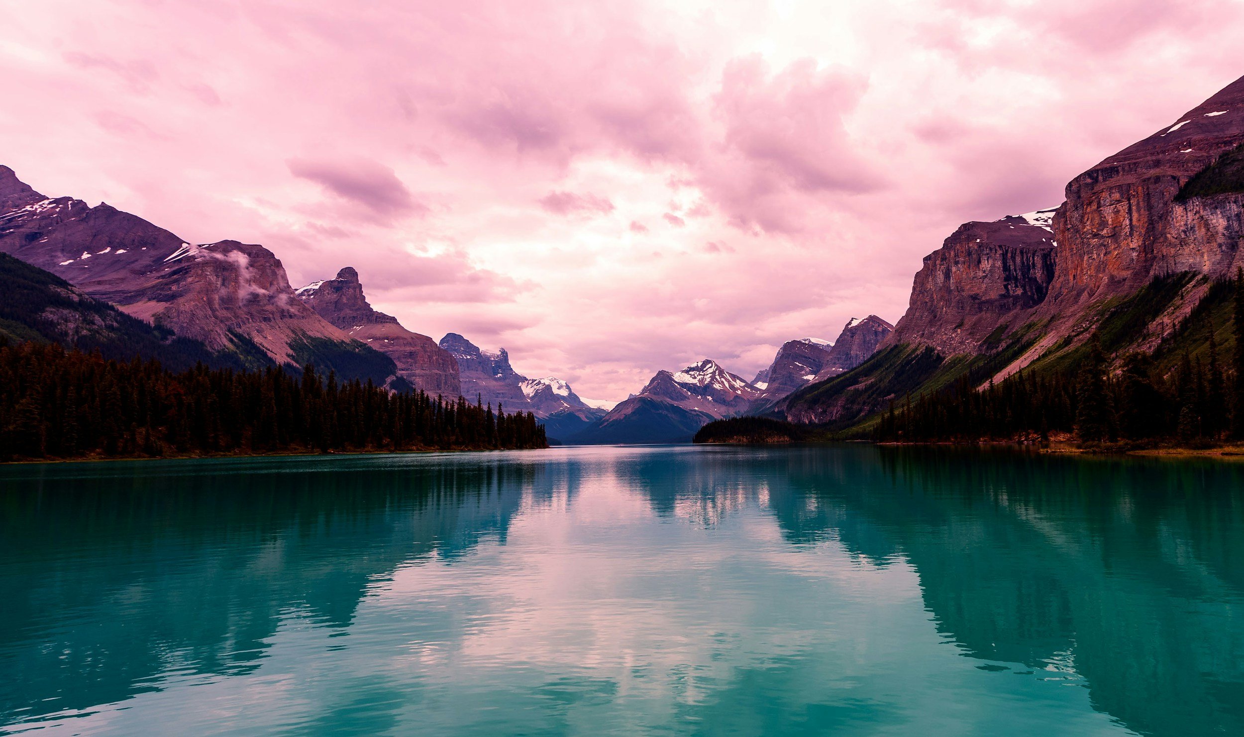 A serene lake reflecting snow-capped mountains and a pink sky with clouds.