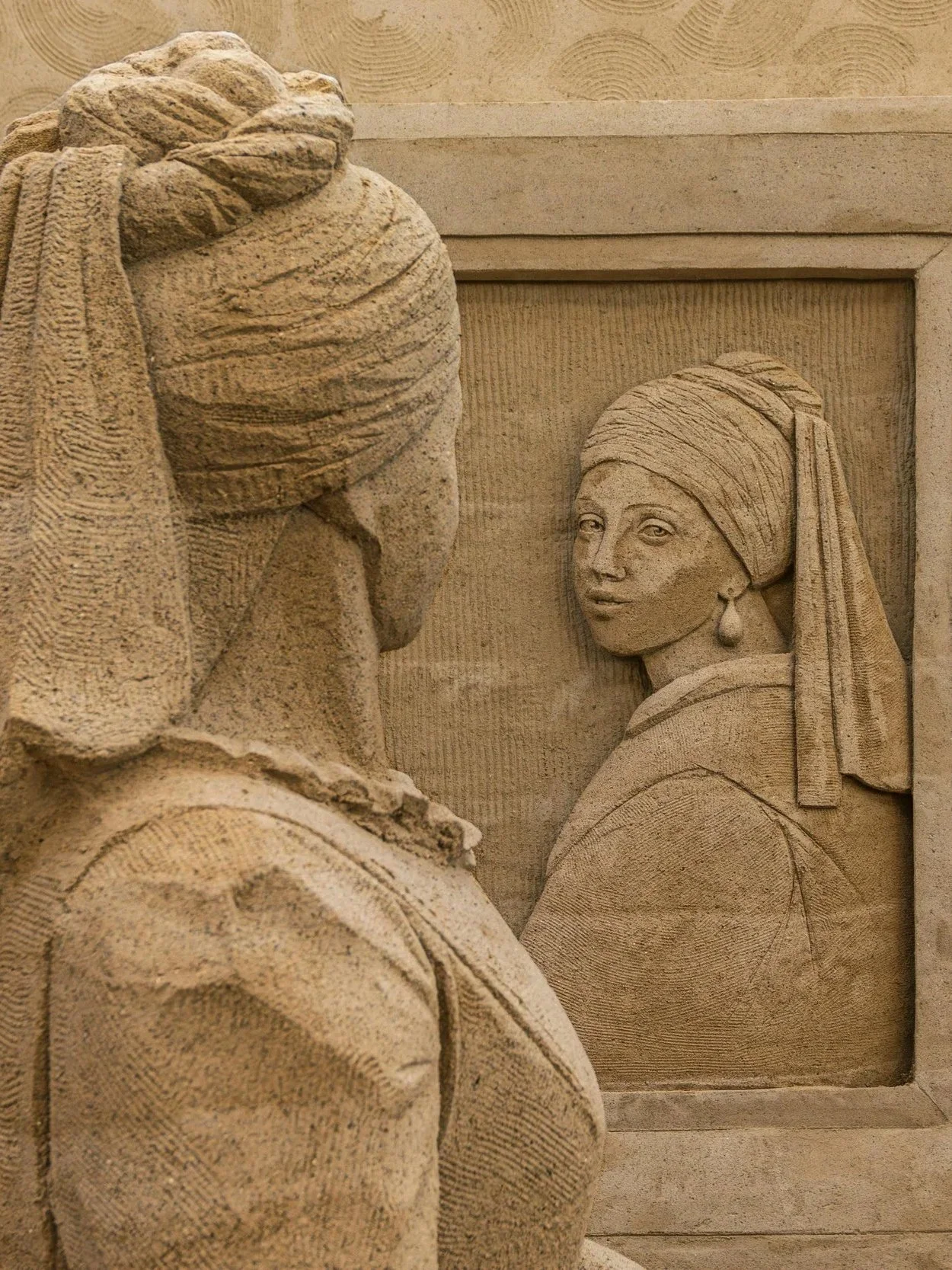 Sandstone relief sculpture featuring a woman with a headscarf and earrings shown in a mirror reflection, with a partially visible back profile of the woman looking at herself.