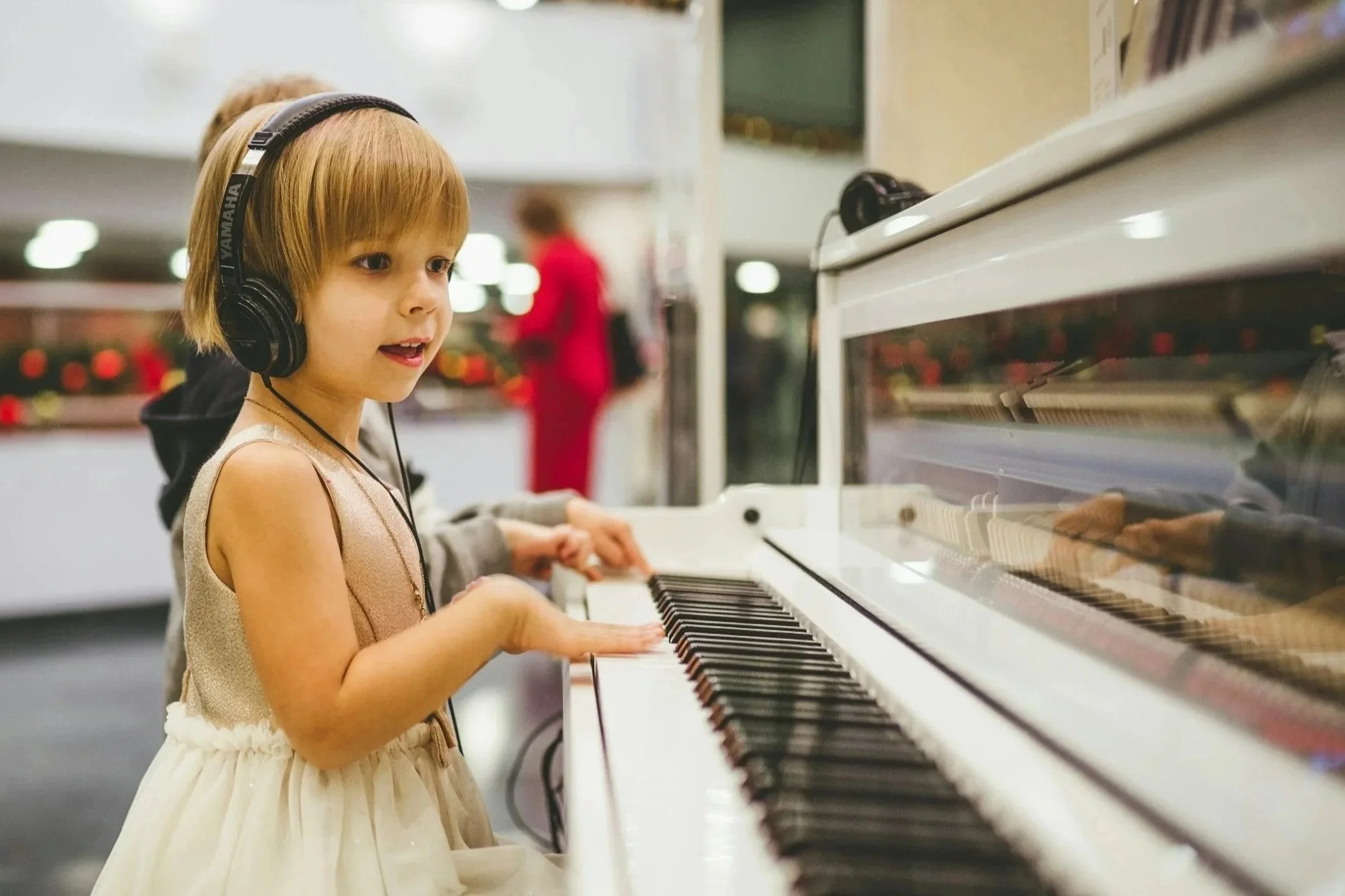 A smiling young child with headphones watches the inner workings of a piano through a clear window on the top of the piano as she plays.