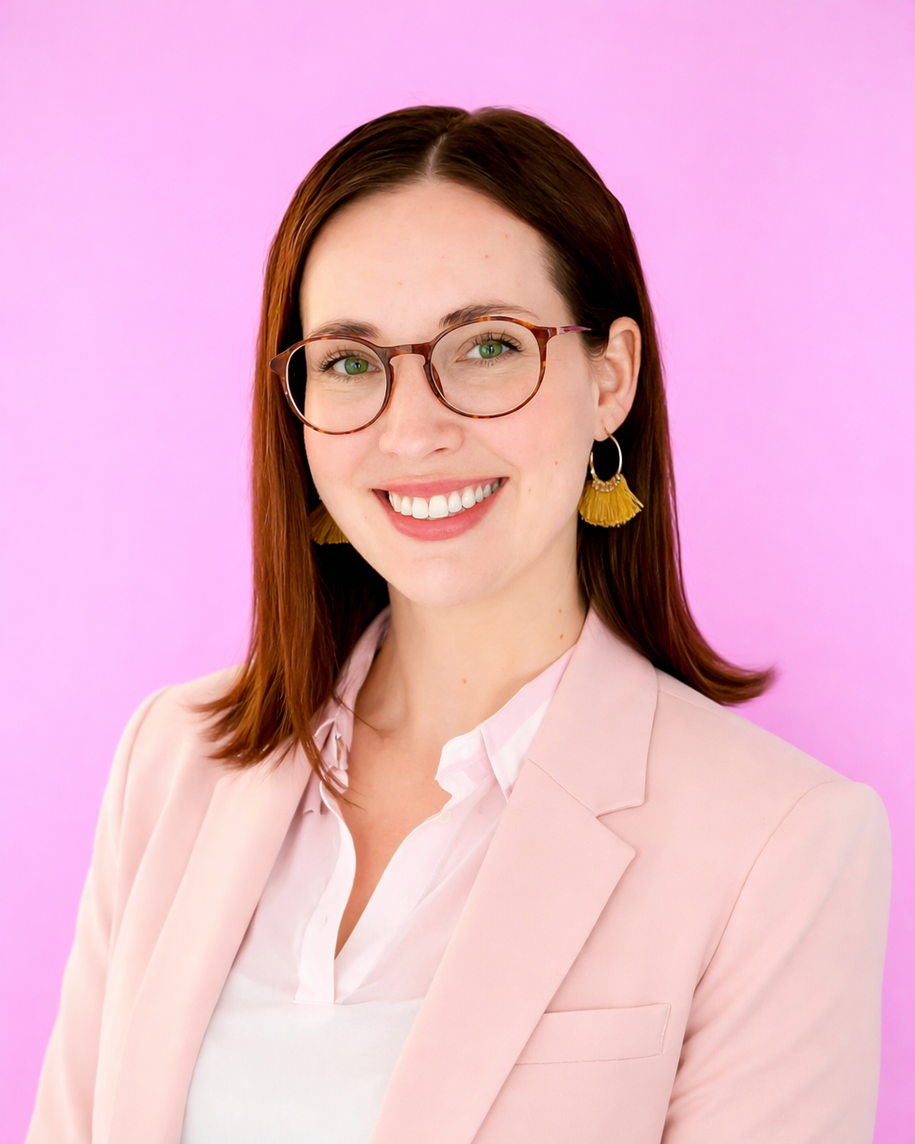 A smiling woman with brown hair, glasses, gold tasseled earrings, wearing a light pink blazer and white and pink striped shirt against a pink background.