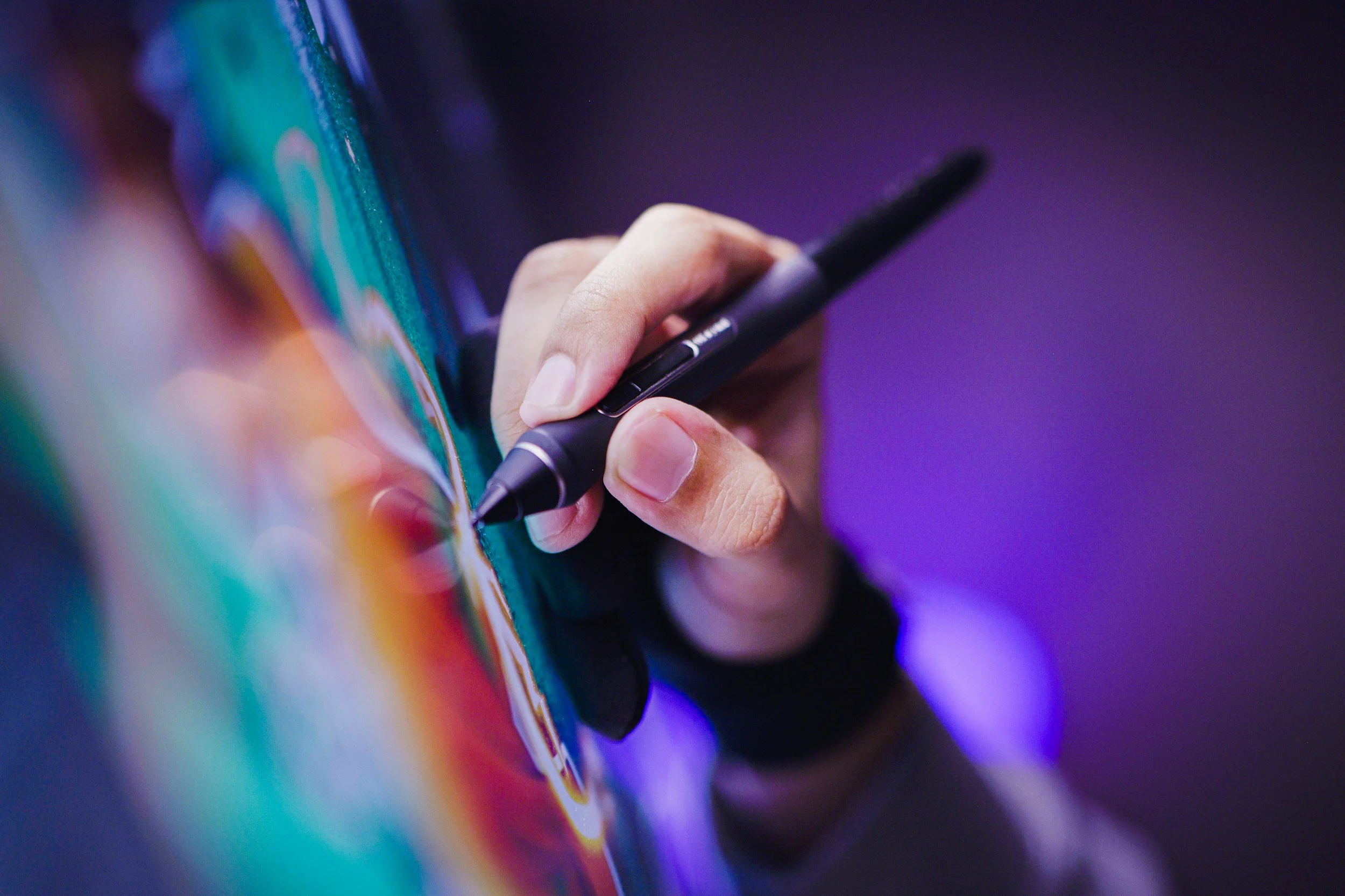 Close-up of a hand holding a stylus pen and drawing on a colorful digital tablet screen with a purple background.