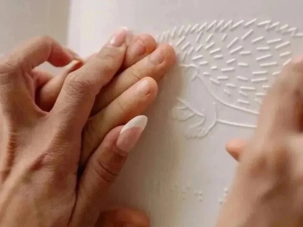 A small hand touches a textured paper with a hedgehog design, with an adult's hands guiding their hands over the paper.