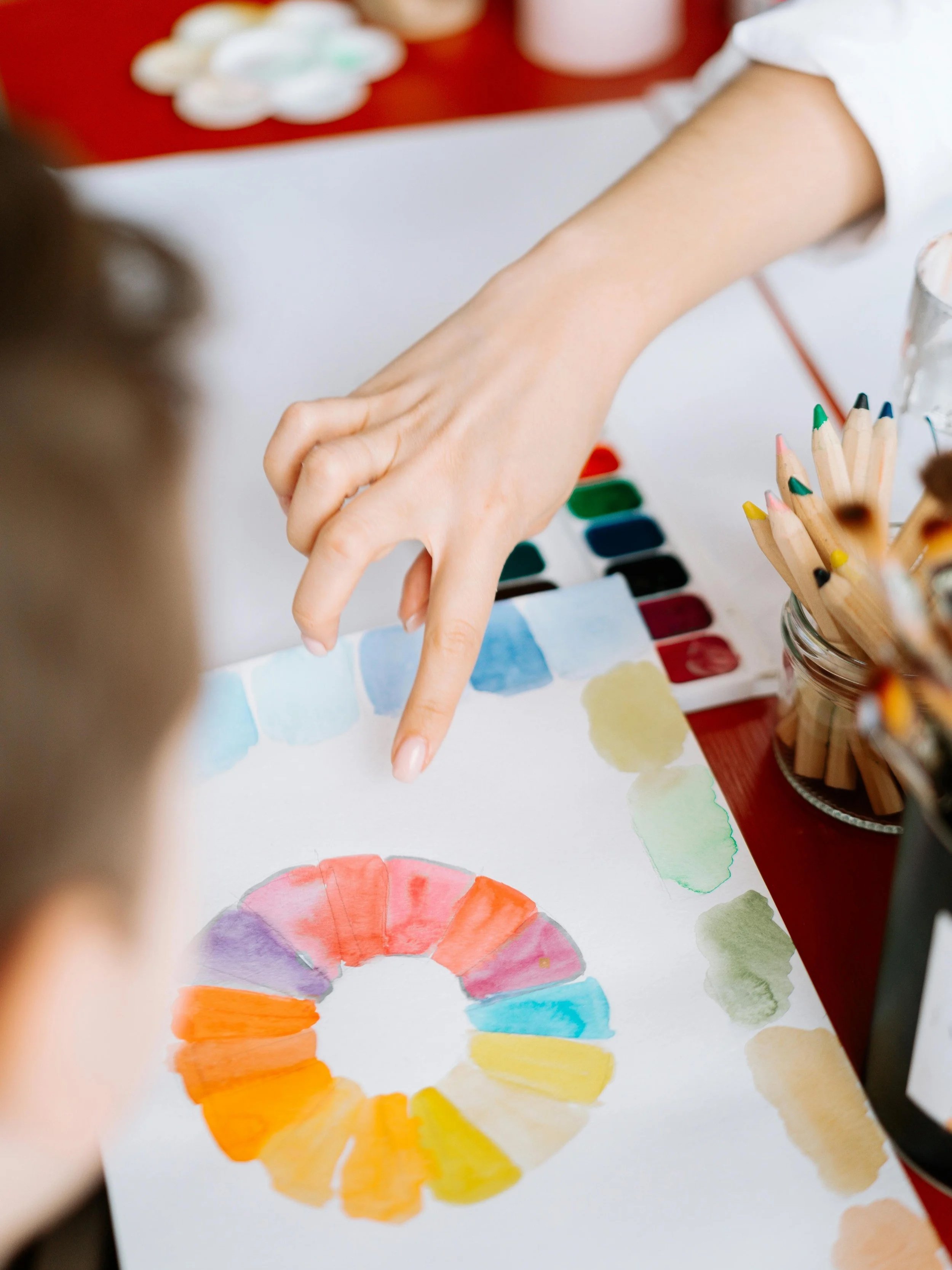 Close-up of a person's hand pointing at a color wheel painted with watercolors on paper, with art supplies such as colored pencils and watercolor paints on a table.