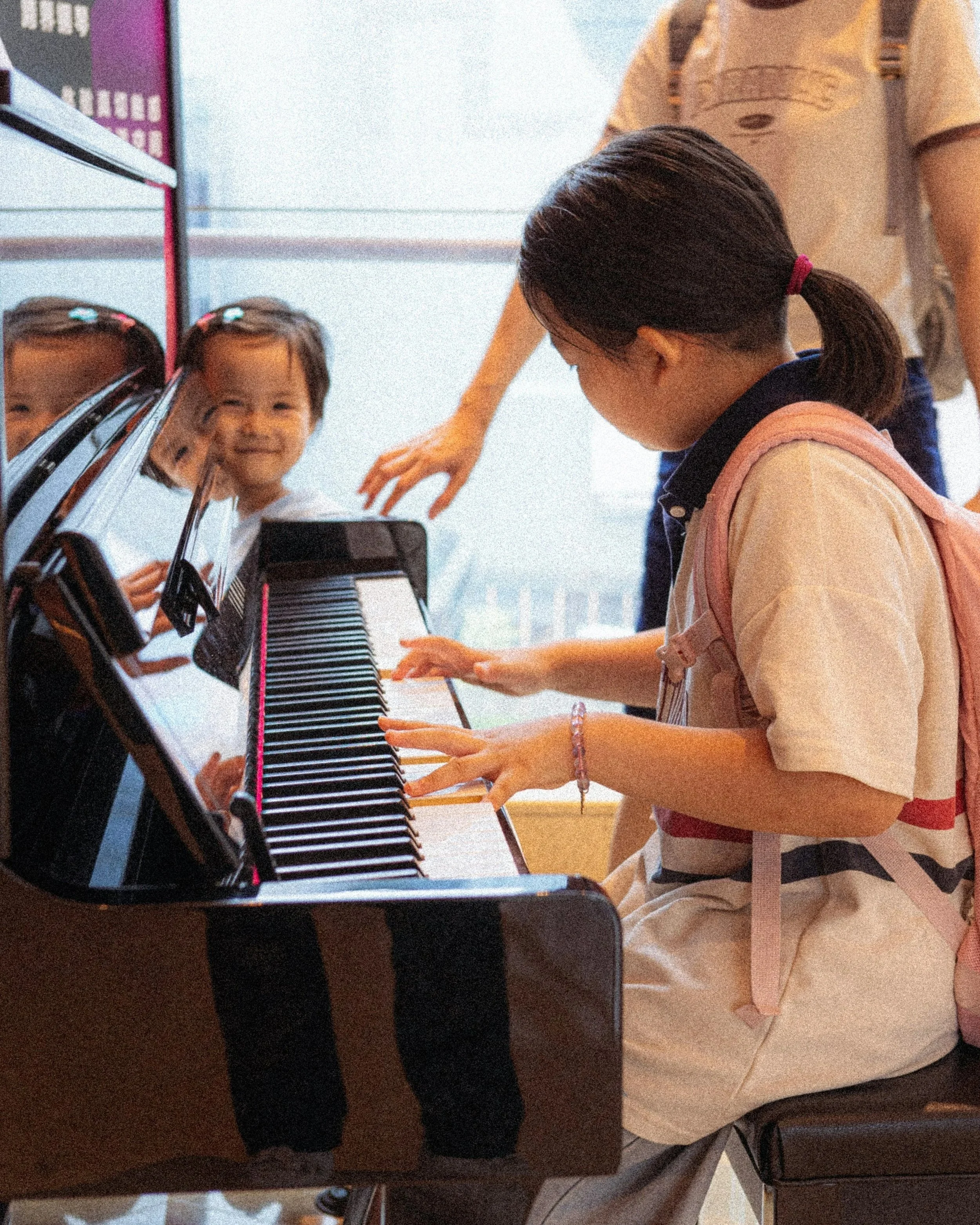 A girl with a ponytail playing the black keys on a piano while looking at the keys, with a peer or teacher standing beside her. A small child smiles as she watches from the side of the piano.