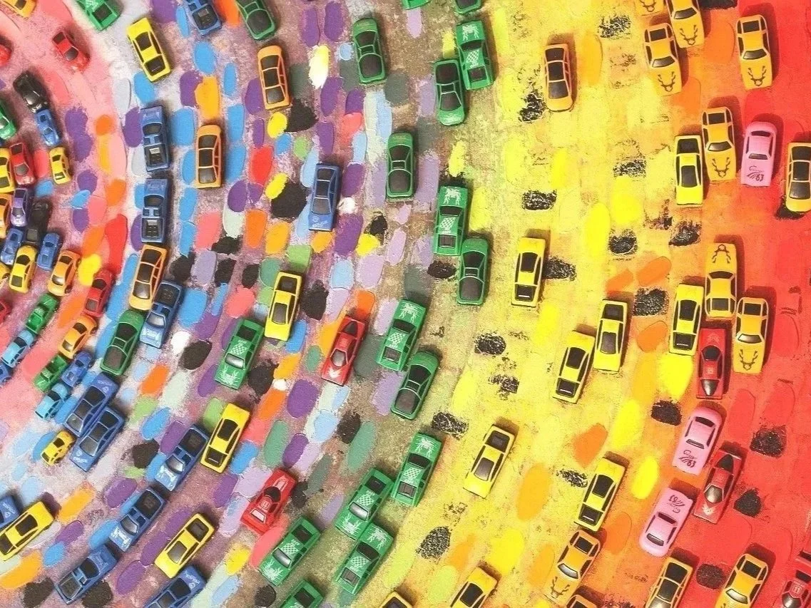 A colorful outdoor art installation with miniature toy cars arranged in rainbow-colored sections on a painted ground resembling a rainbow.