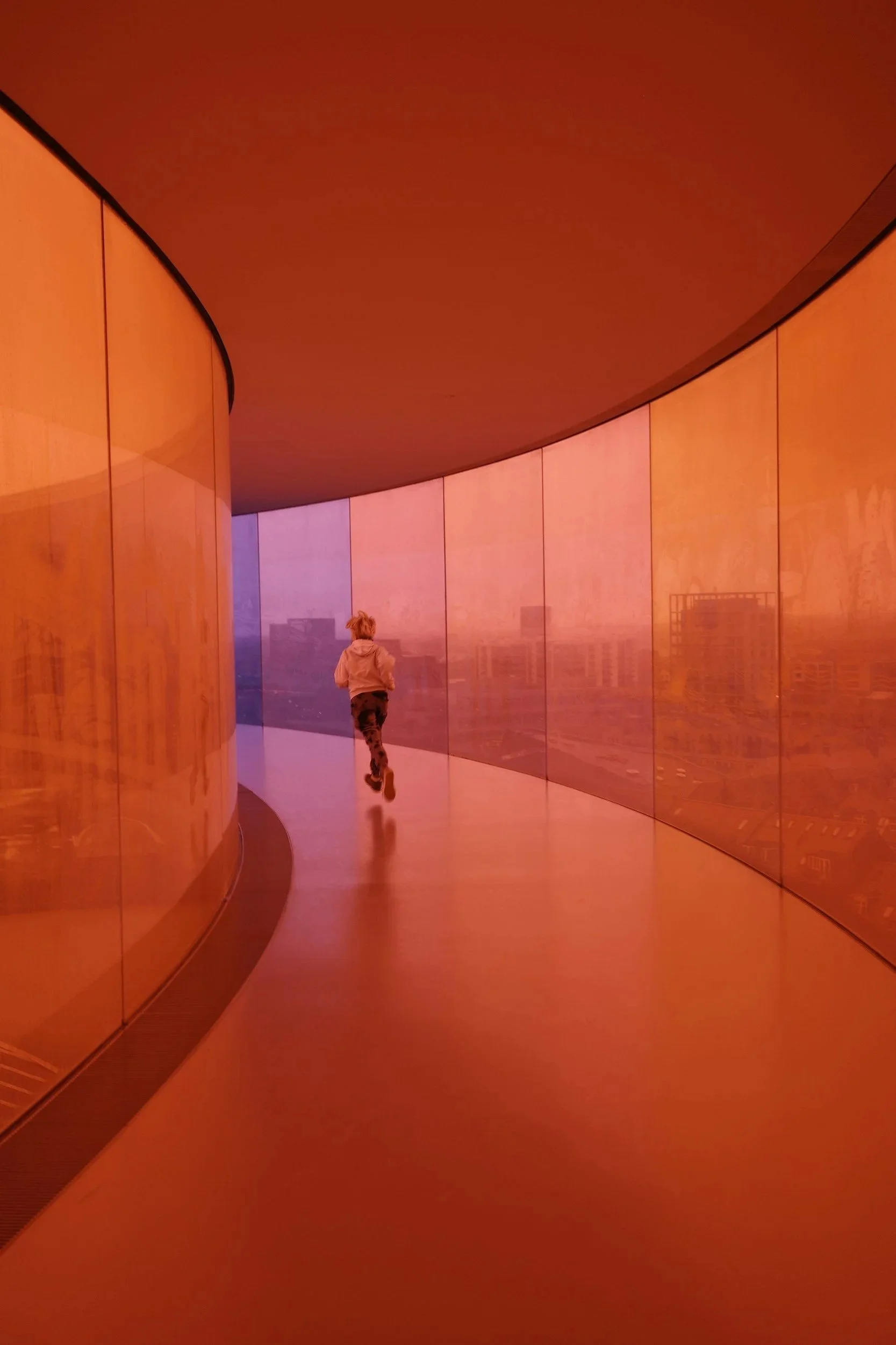 A child running in a curved, orange and purple-lit corridor with reflective walls and windows with a cityscape view in the background.