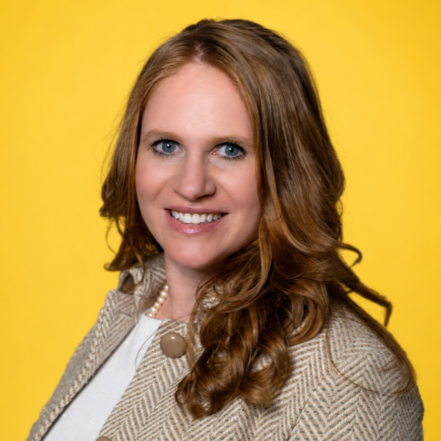 A smiling woman with red hair, a white shirt, a light tan tweed jacket, and a pearl necklace with a marigold colored background.