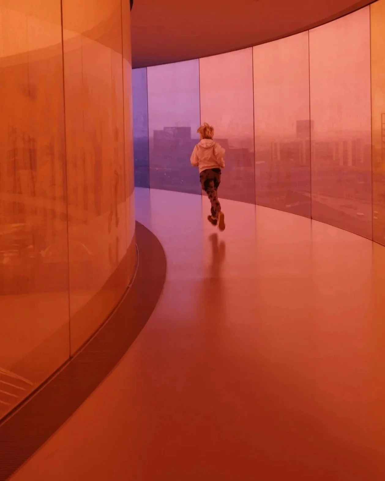 A child runs along a curved glass hallway with orange-tinted walls and purple-tinted glass panels, city skyline visible through the glass.