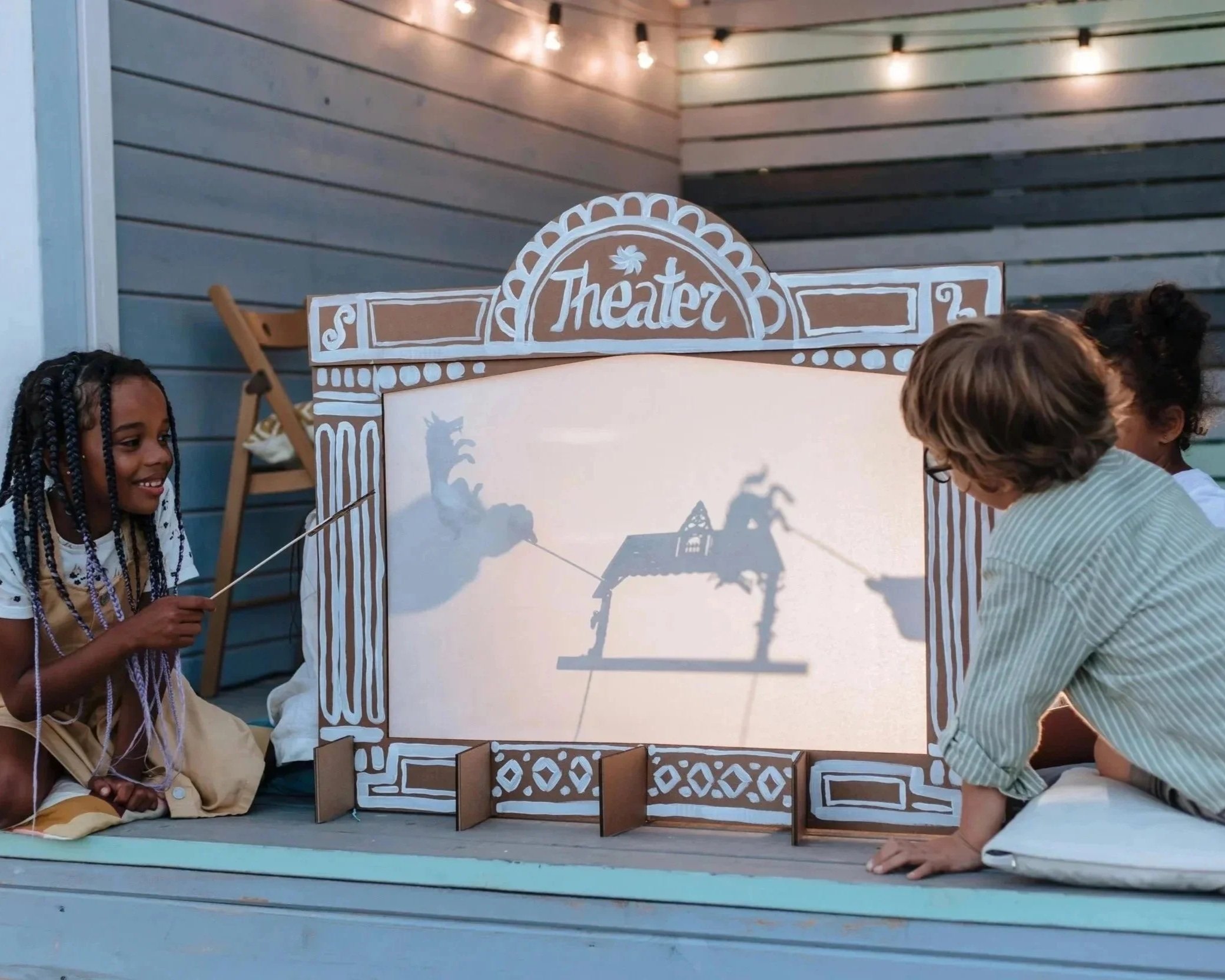 Children performing shadow puppetry in front of a small theater stage with a white backdrop and a wooden frame labeled 'Theater' at the top.