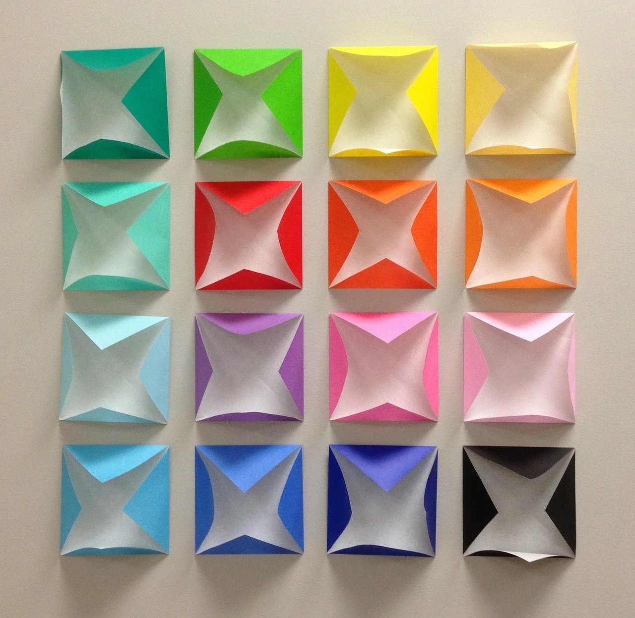 Colorful origami squares arranged in a 4x4 grid on a white wall, with each square featuring a pyramid-like shape in various colors including teal, green, yellow, orange, pink, purple, blue, and black.