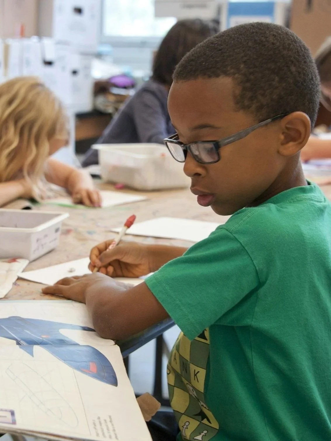 Two children are lying on a table, closely examining a colorful picture in an open book. One child is holding a pen and pointing at the picture.