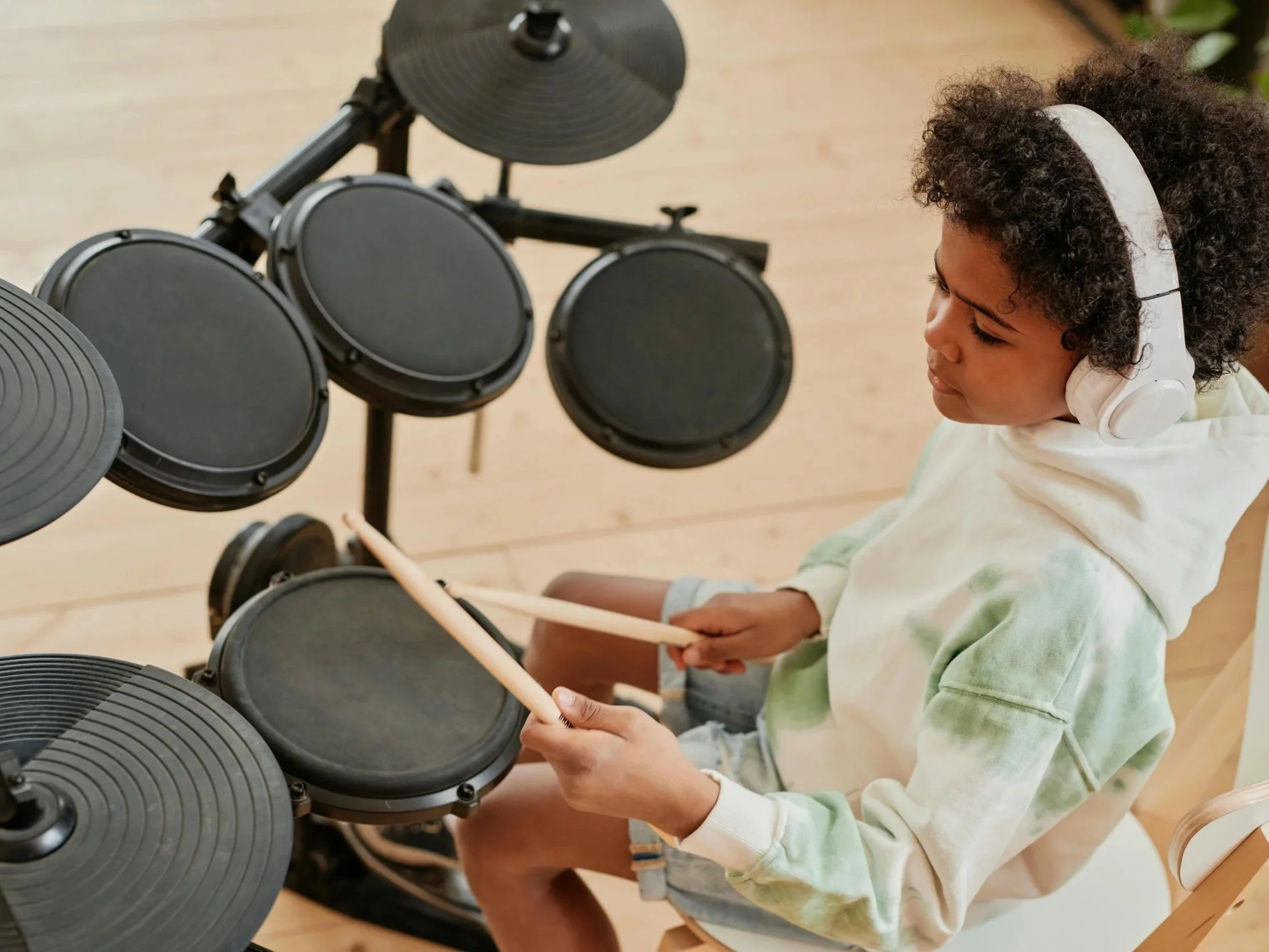 A young girl wearing headphones plays electronic drum pads with drumsticks.