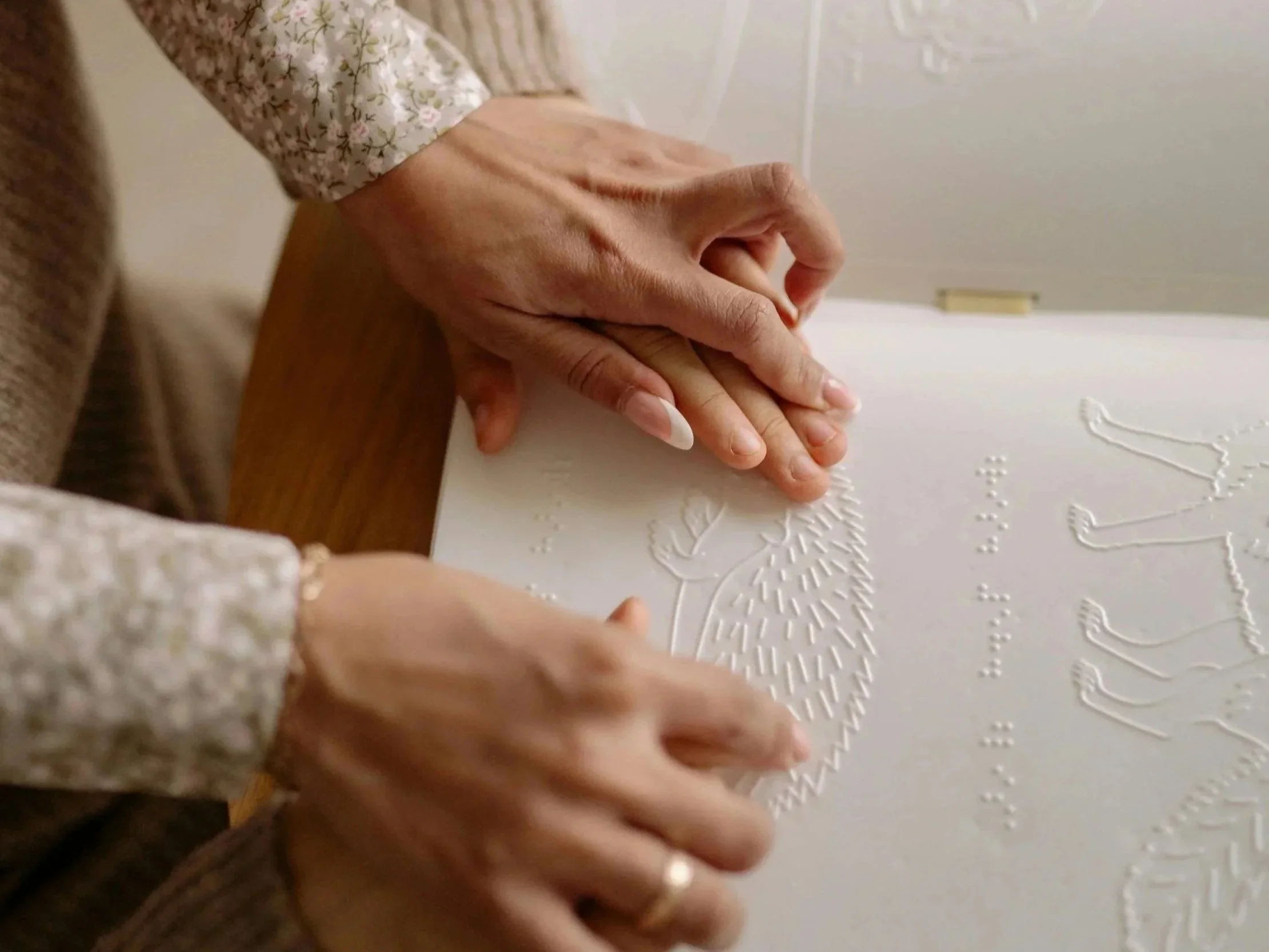 An adult guides the hands of a child over a raised textured image of a hedgehog and braille.