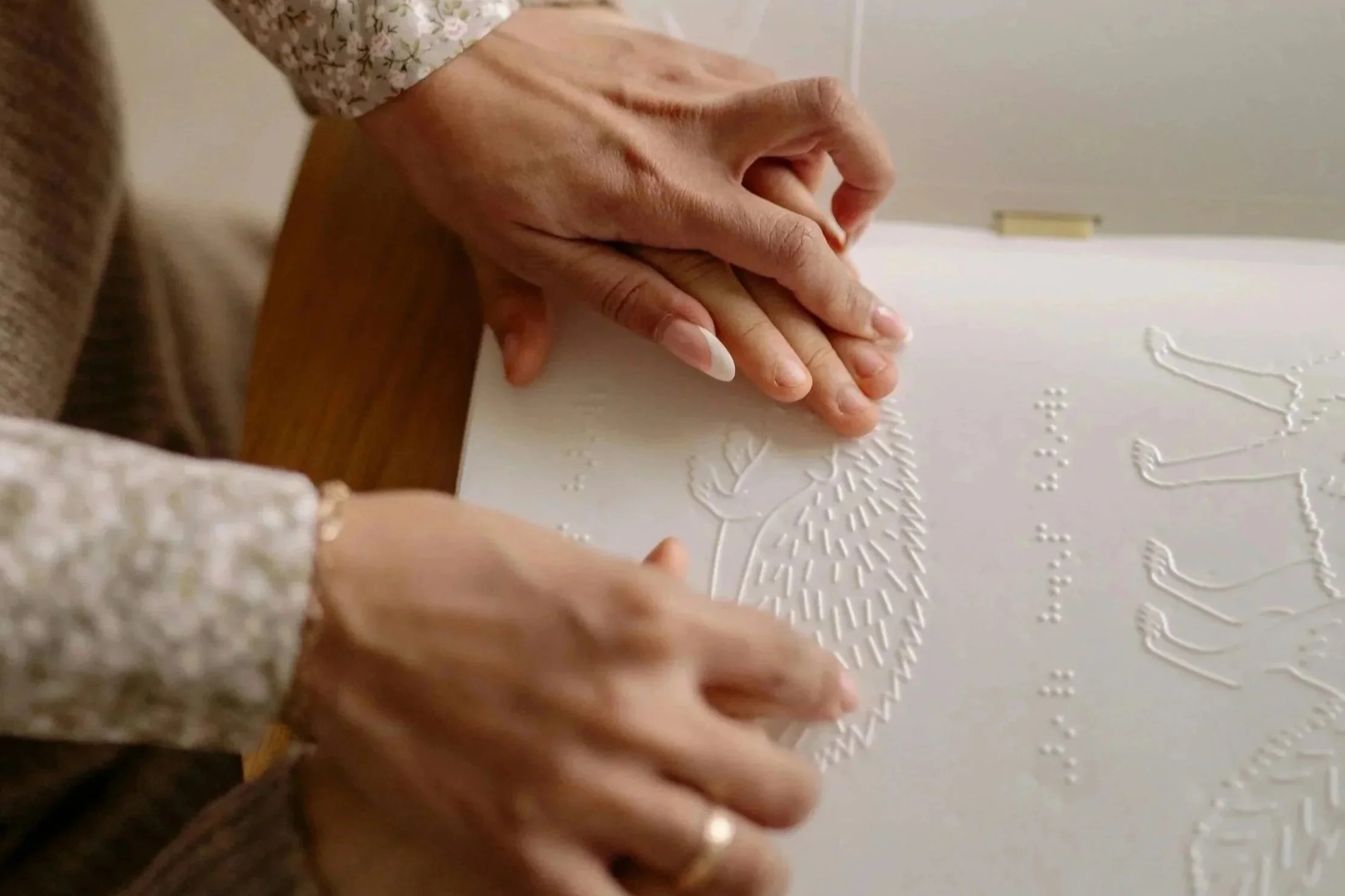 A parent guides the hands of a child over a raised line drawing of a hedgehog and braille.