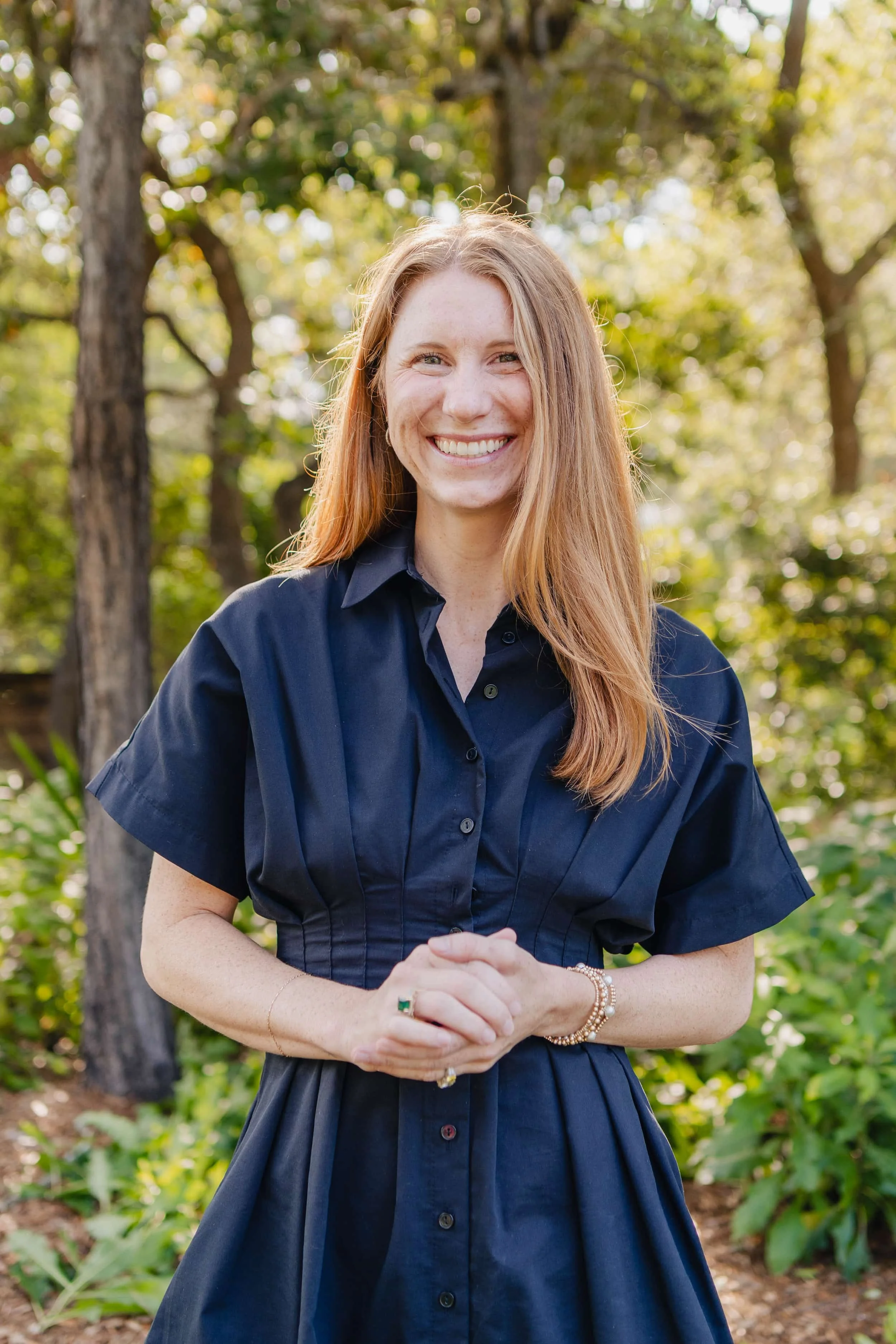 A woman with long red hair wearing a navy blue dress, standing outdoors in a wooded area, smiling at the camera.