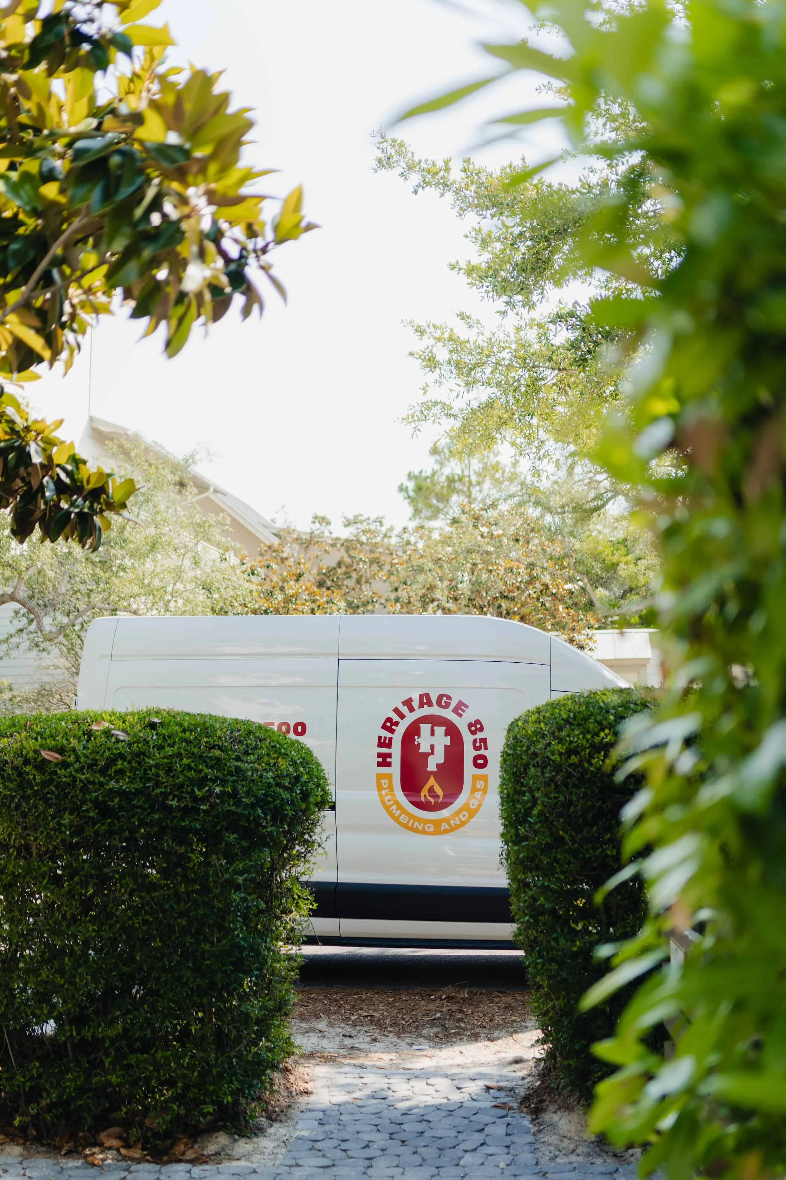 A white Heritage Plumbing and Gas service van parks behind a hedge, partially visible through a gap in the greenery, on a sunny day.