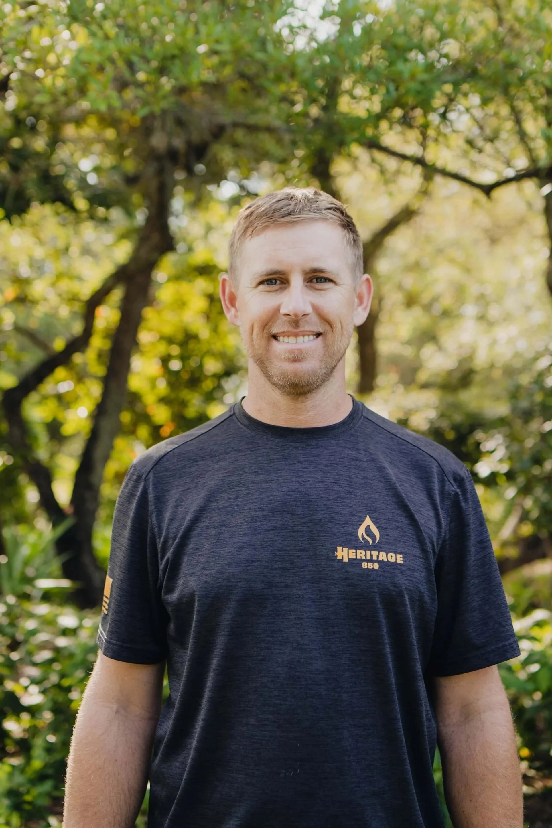 A smiling man outdoors, standing in a park or woods with green trees and foliage in the background. He is wearing a black t-shirt with a gold logo and text that reads 'HERITAGE 850' and a flame symbol.