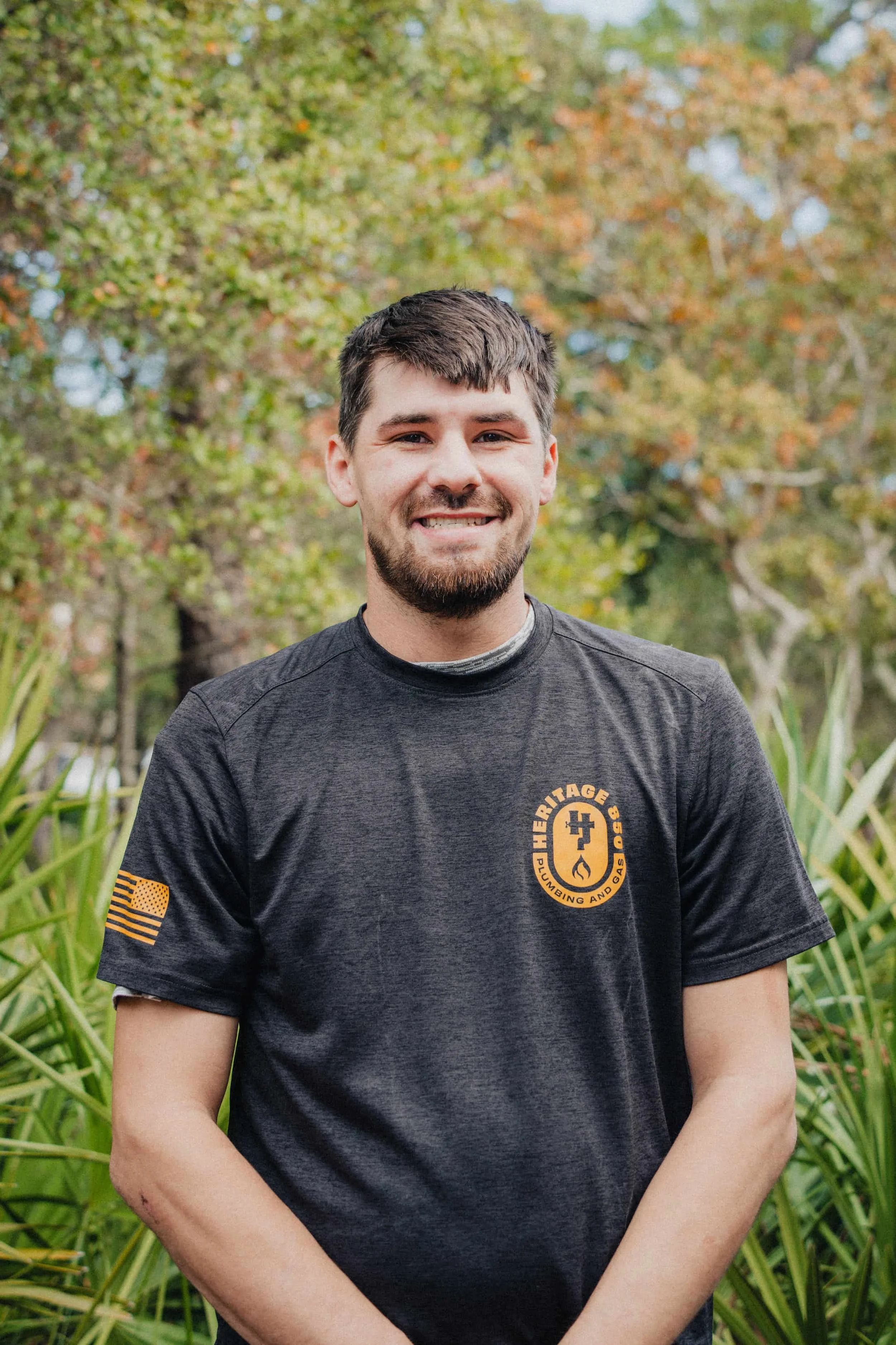 A smiling man standing outdoors in front of green plants and trees, wearing a black t-shirt with a logo that says 'Heritage Plumbing and Gas' and an American flag patch on his sleeve.