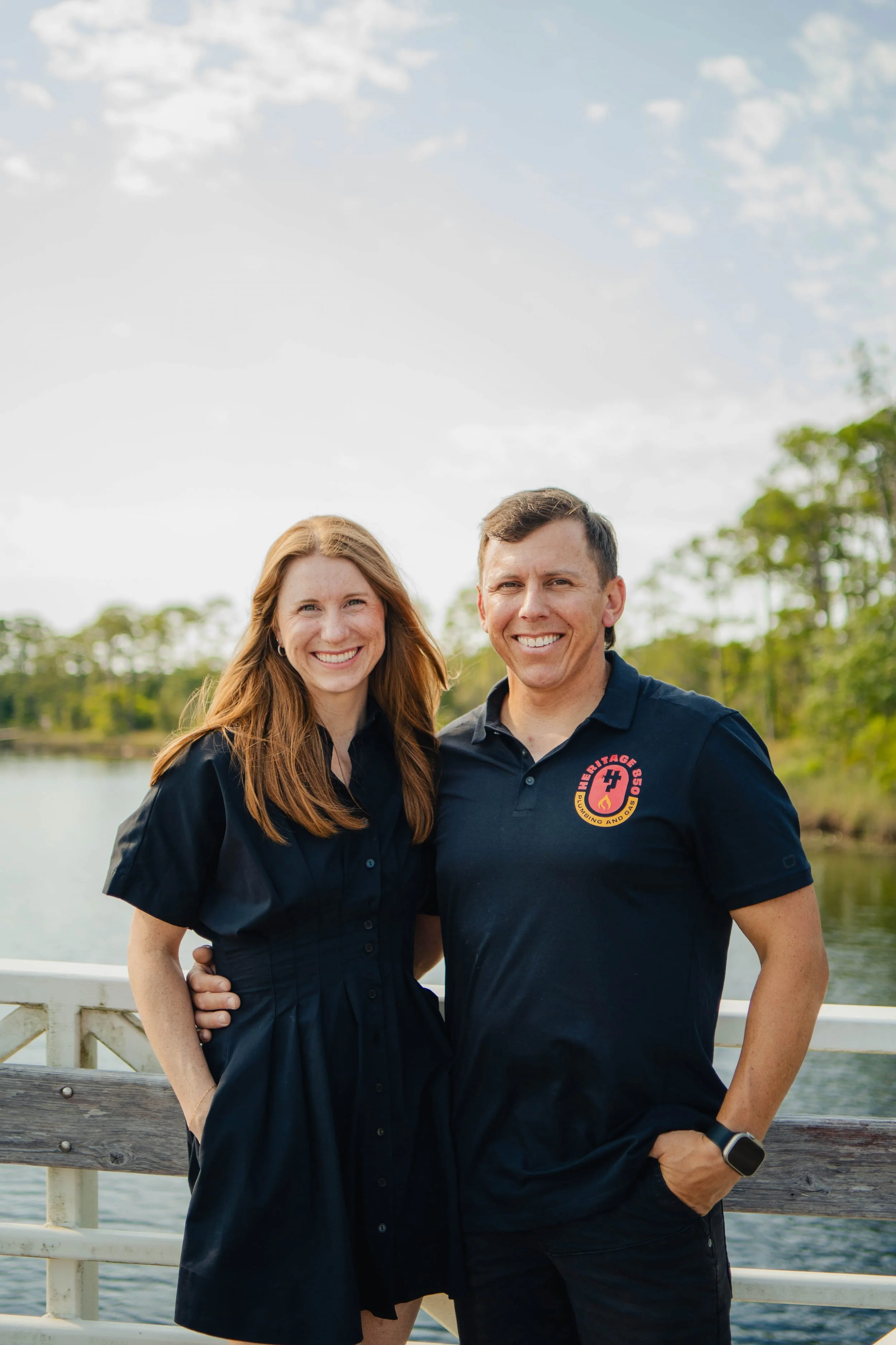 A smiling couple standing outdoors on a bridge near water with trees in the background, both wearing dark shirts, the woman's dress is buttoned down, and the man's shirt has a logo on it.