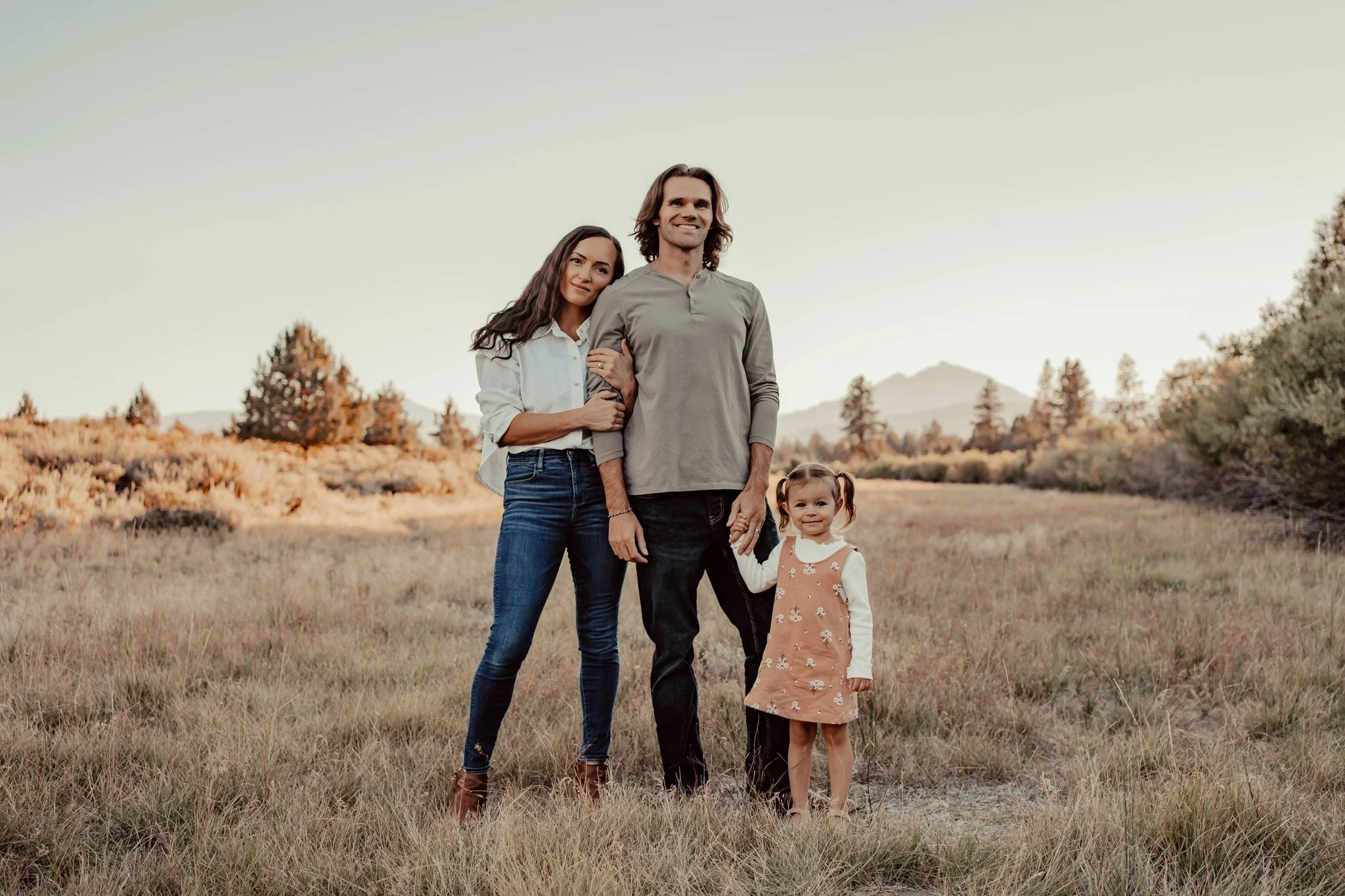 A family of three standing outdoors in a grassy field with trees and mountains in the background during sunset or sunrise.