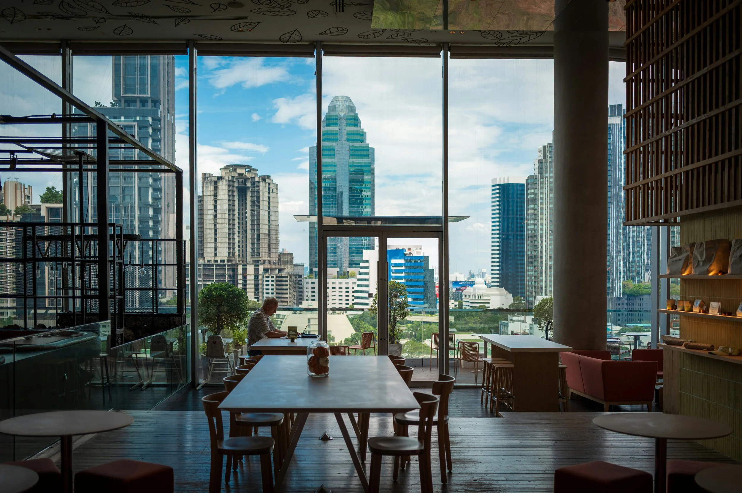Interior of a modern cafe with a person sitting at a table by large glass windows overlooking a city skyline with various tall buildings on a bright day.