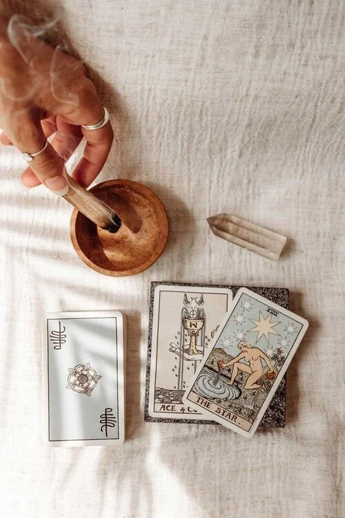 A hand holding a smoked stick over a wooden dish, with tarot cards and a crystal nearby on a light-colored cloth surface.