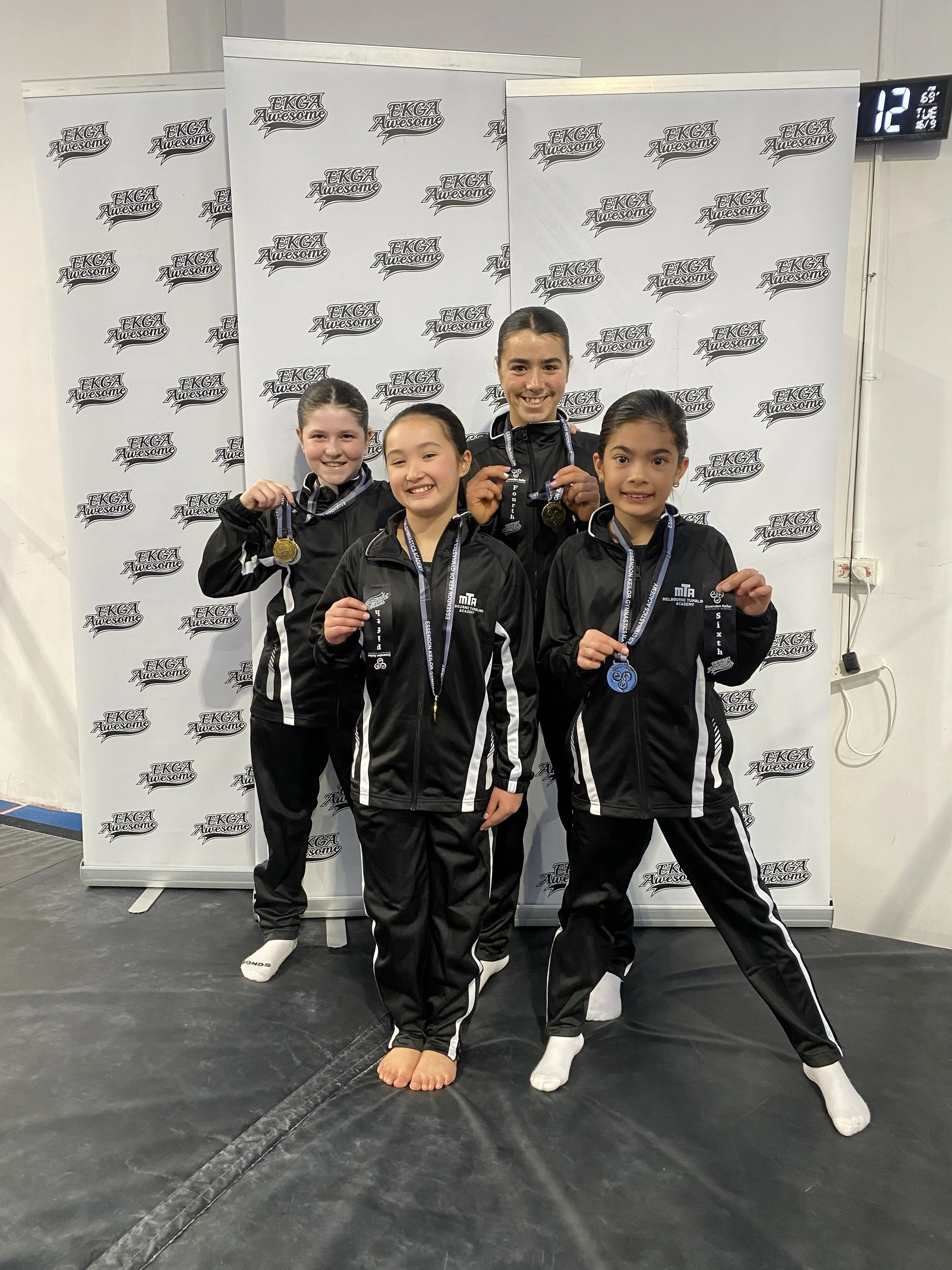 Four young female gymnasts in black tracksuits with white stripes, standing on a podium with medals around their necks, smiling after a competition, with a backdrop that reads 'EKGA Awesome'.