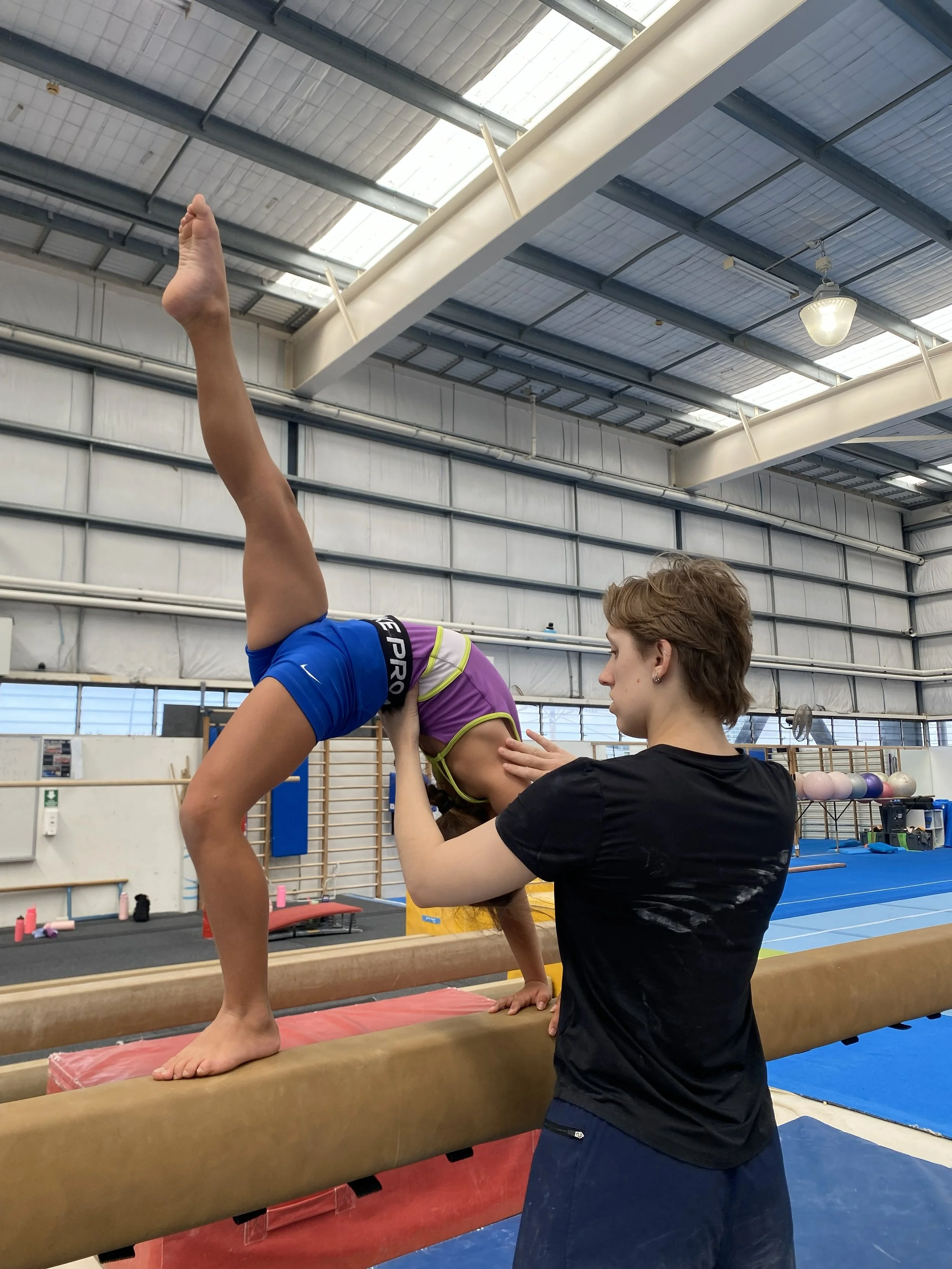 A young female gymnast practicing handstands on a balance beam in a gymnasium, guided by a coach.