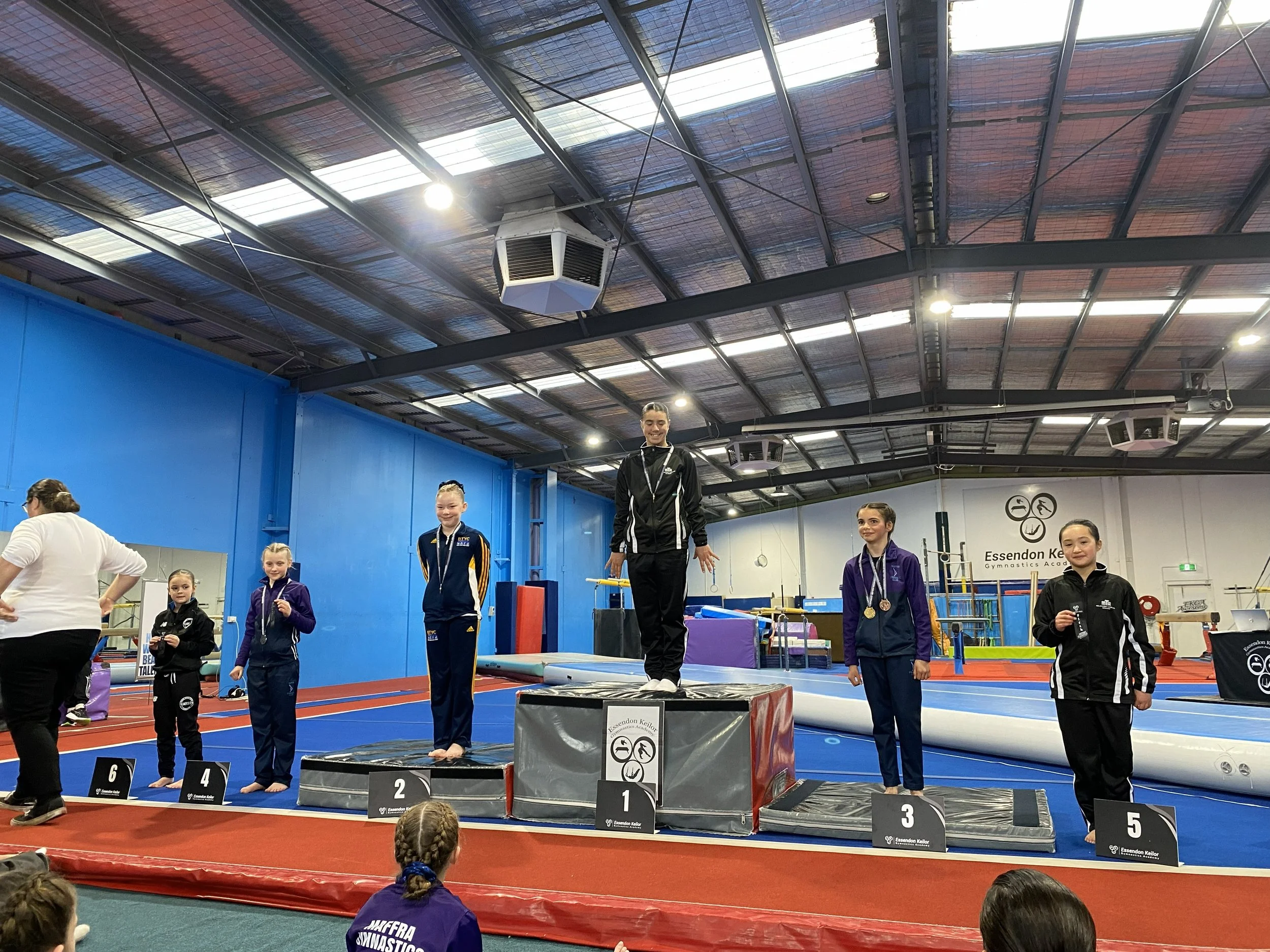 Award ceremony at a gymnastics competition, with young gymnasts standing on a podium receiving medals, in an indoor gymnasium.