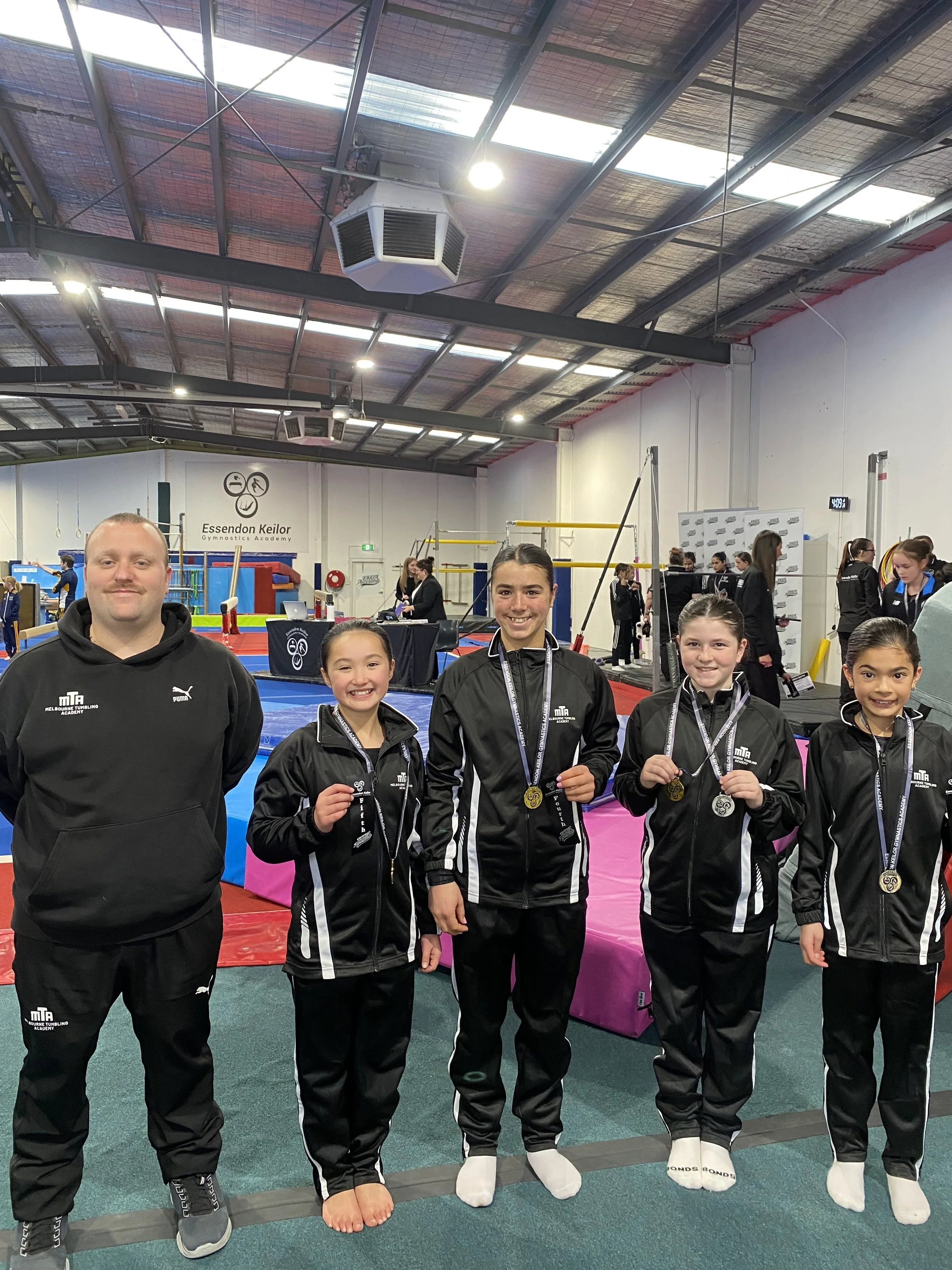 Group of five people, with one adult and four young girls, standing inside a gymnastics facility, smiling, wearing black tracksuits, and medals around their necks.